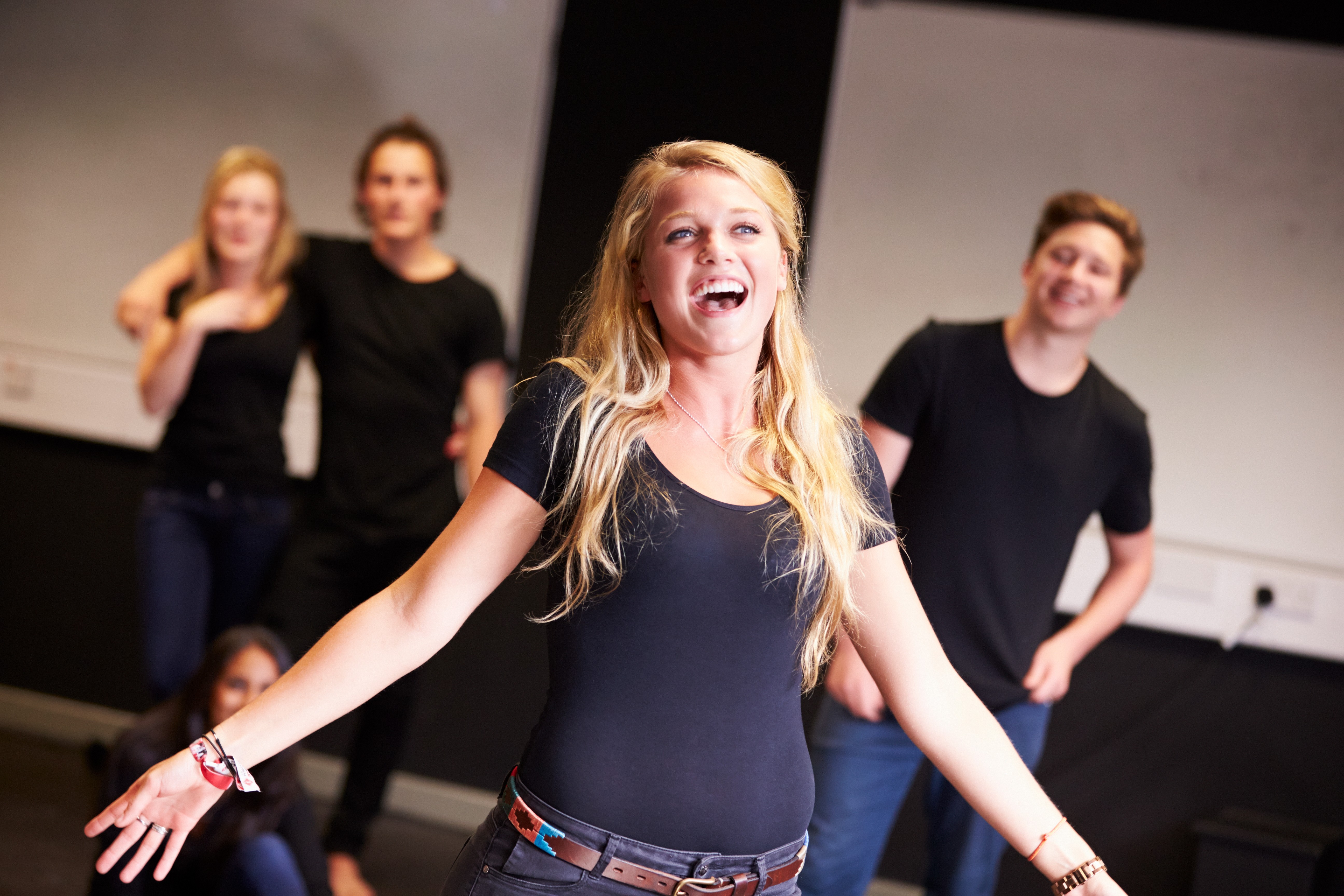 A blonde woman smiling brightly on stage with her arms outstretched, dressed in a black outfit, with a spotlight behind her.