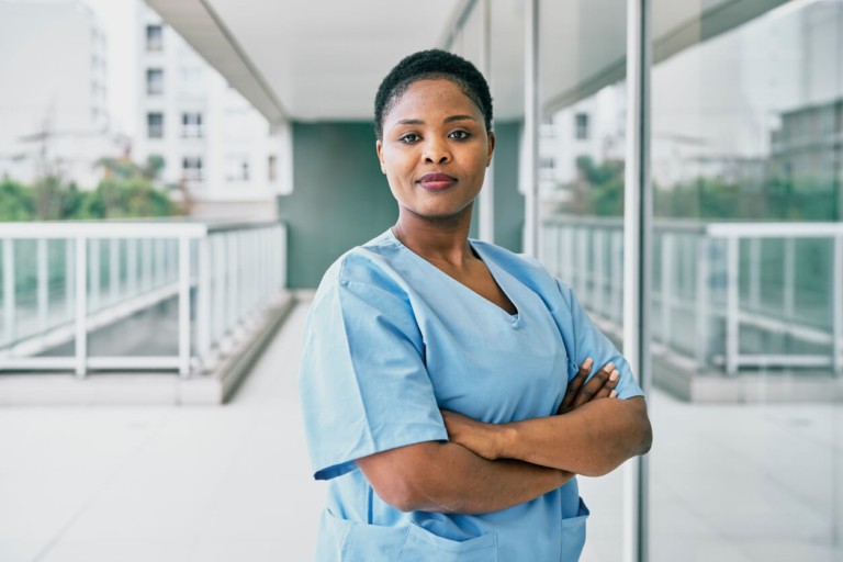 A nurse in blue scrubs standing with arms crossed in a healthcare setting, looking directly at the camera
