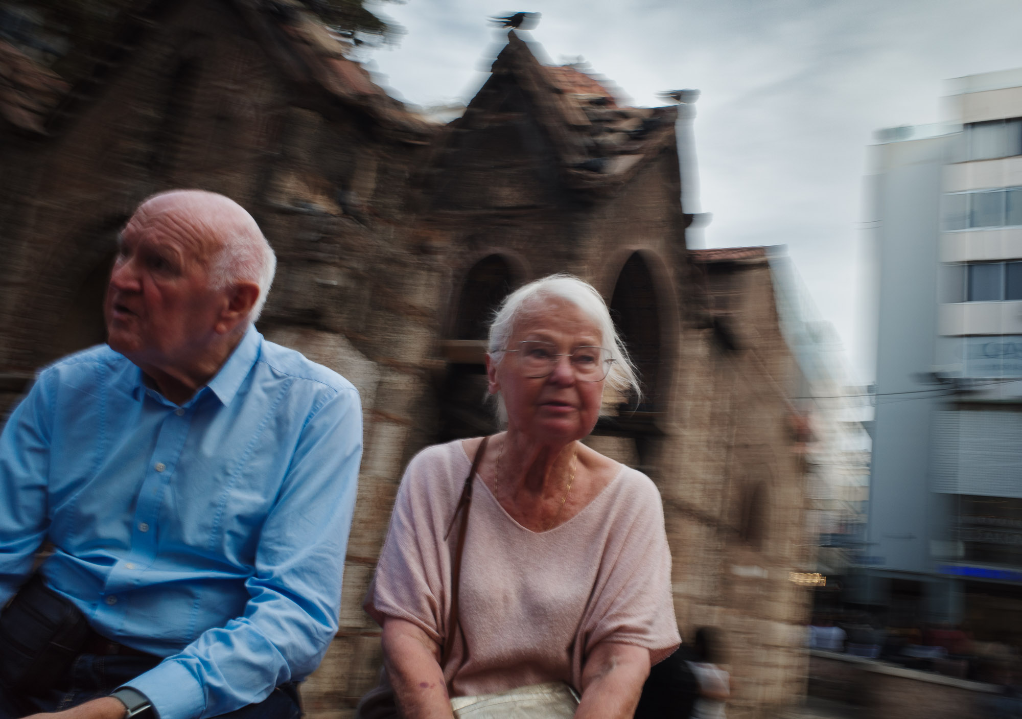 An elderly couple sits together on a bench in front of a rustic, stone building, with a blurred, cityscape backdrop under a cloudy sky.