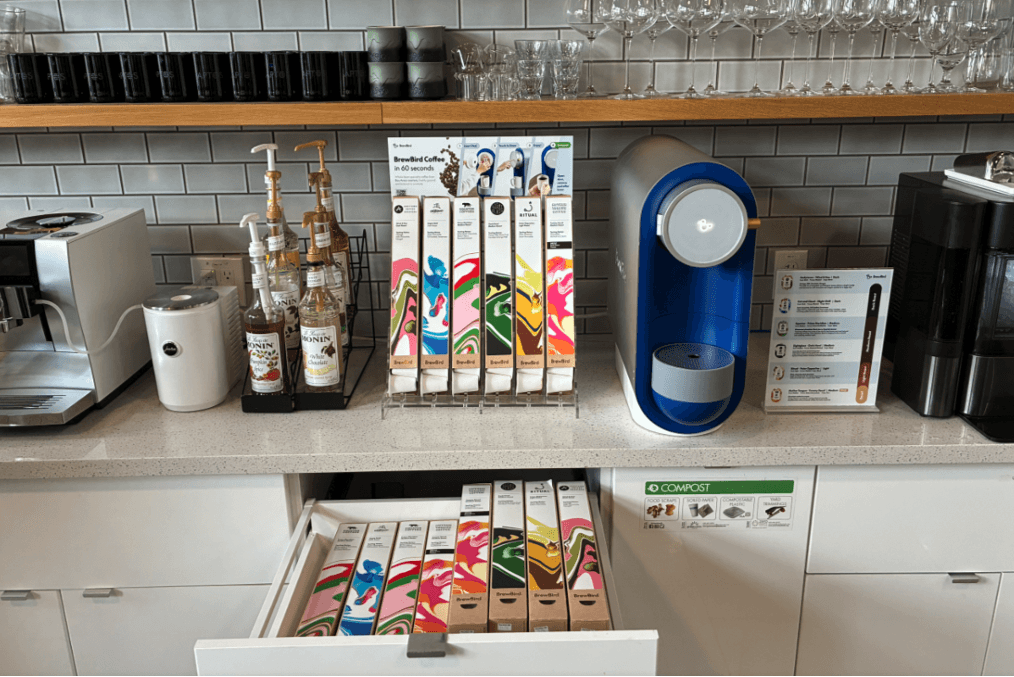Title: BrewBird integrated into a mixed coffee station - Description: A BrewBird brewer on a counter with syrups, coffee storage, and other office coffee equipment.