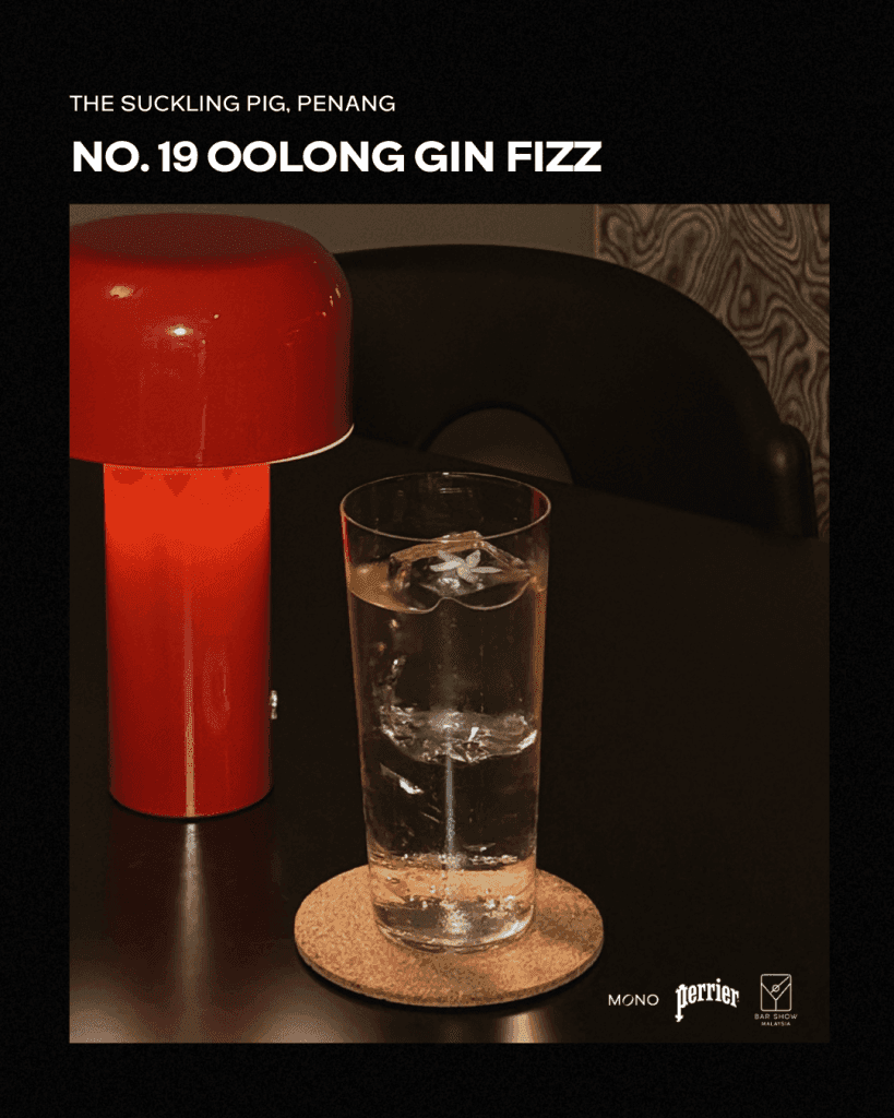 A tall glass of Oolong Gin Fizz with ice on a cork coaster, accompanied by a red lamp in the background, displayed on a dark table.