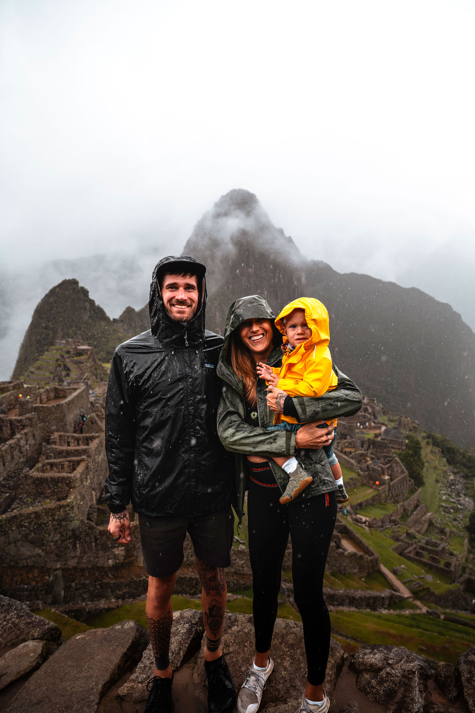 Bre and Flo smiling wearing raincoats stands in front of misty mountains at Machu Picchu.
