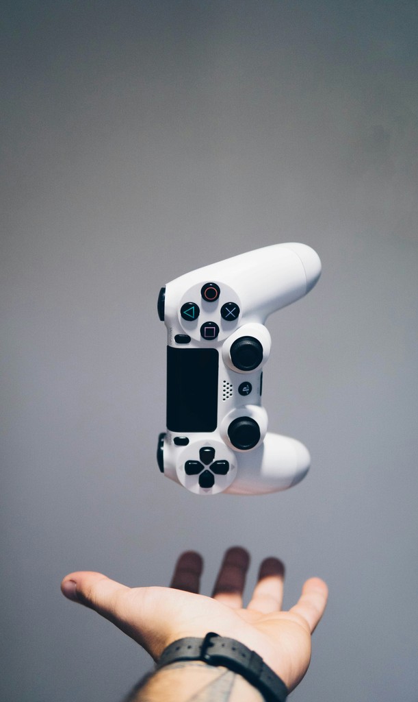 A hand with a wristwatch appears to toss a white game controller in mid-air against a plain grey background. The suspended controller emphasizes the dynamic and exciting nature of gaming, showcasing the anticipation and engagement that comes with interactive entertainment.