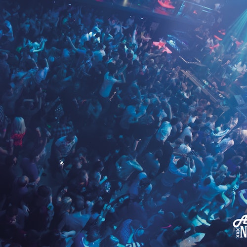 熙攘的夜总会舞池，人们在蓝色和紫色灯光下跳舞，背景中可以看到DJ台。