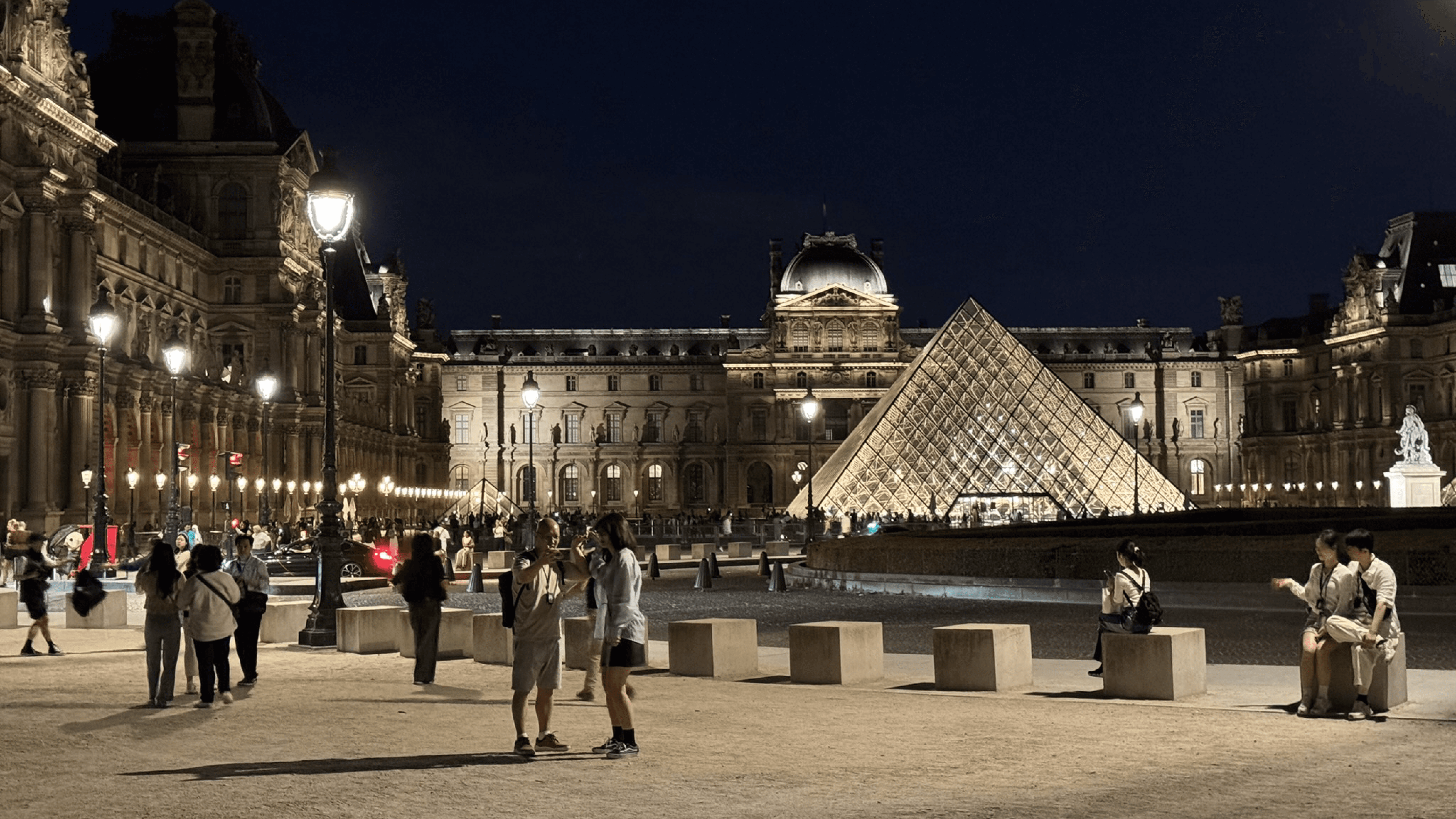 Night view of the Louvre Museum in Paris with the illuminated glass pyramid surrounded by historic palace architecture and visitors in the courtyard.