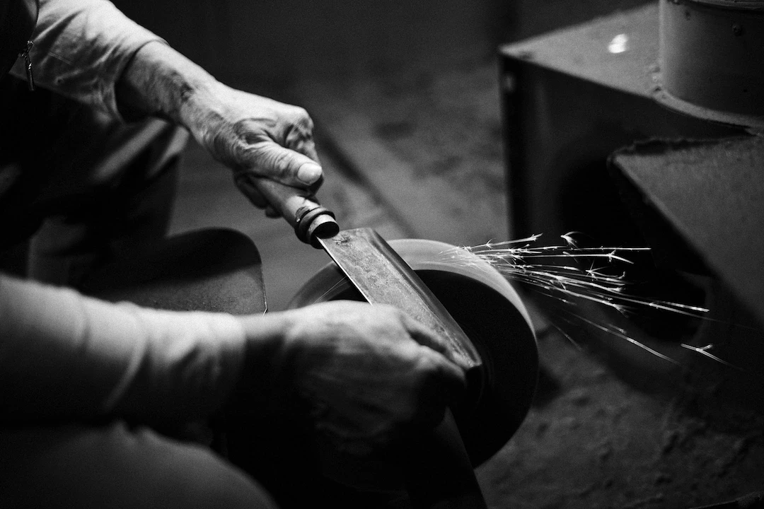 Hands are holding a knife horizontally with the edge touching a spinning sharpening stone.