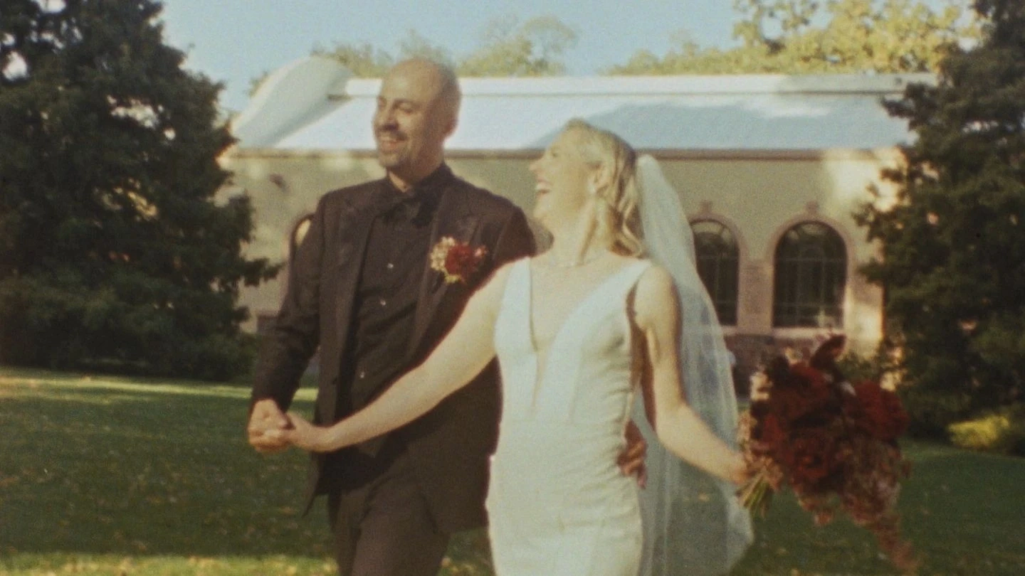 A joyful couple holds hands and smiles under bright sunlight in a lush garden setting, with the bride wearing a white gown and veil, and the groom in a dark suit, framed by a historic building with arched windows in the background, emphasizing the elegance and joy of their wedding day.