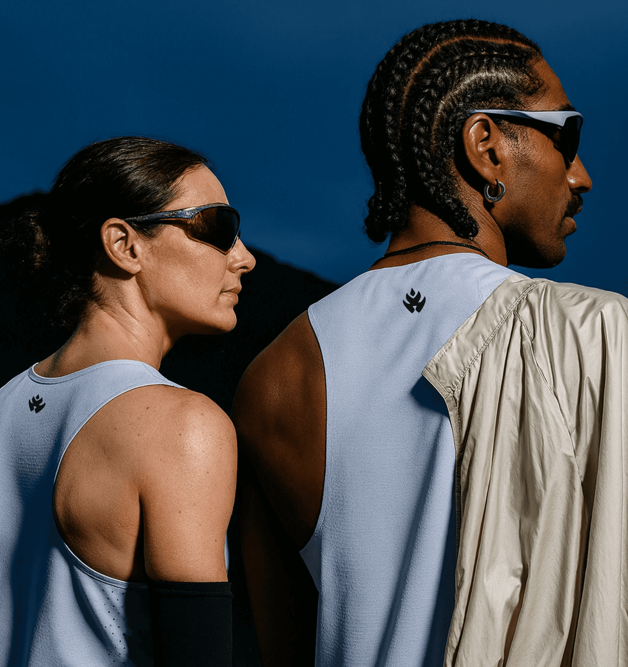 Two athletes wearing light gray sports tanks and sunglasses, standing side by side with their backs facing the camera against a dark sky.