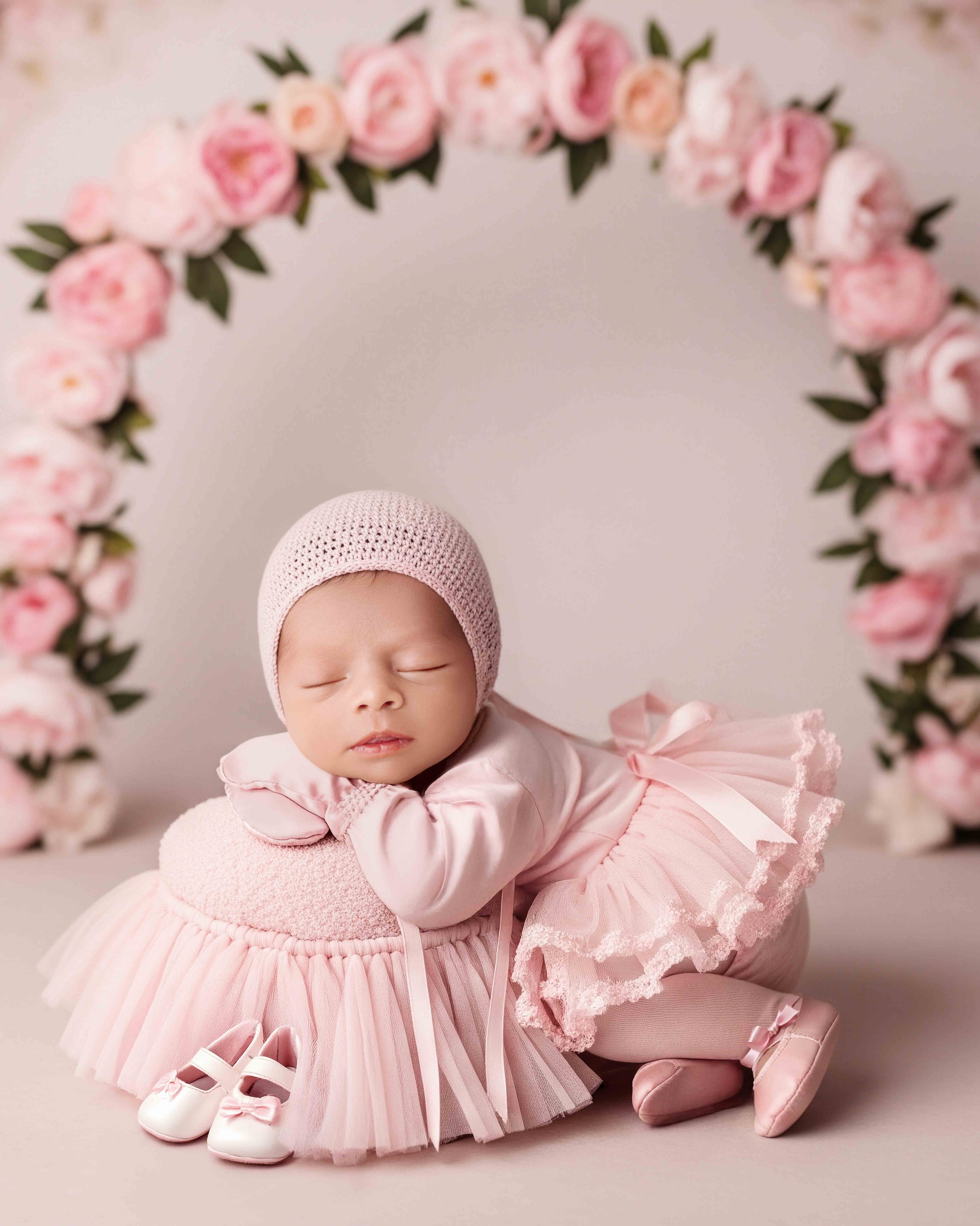 Newborn baby girl in soft pink outfit surrounded by floral arch during studio photoshoot