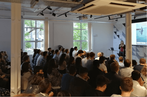 An event with a room of people facing a speaker (Samantha) who is standing at the front next to a projection..