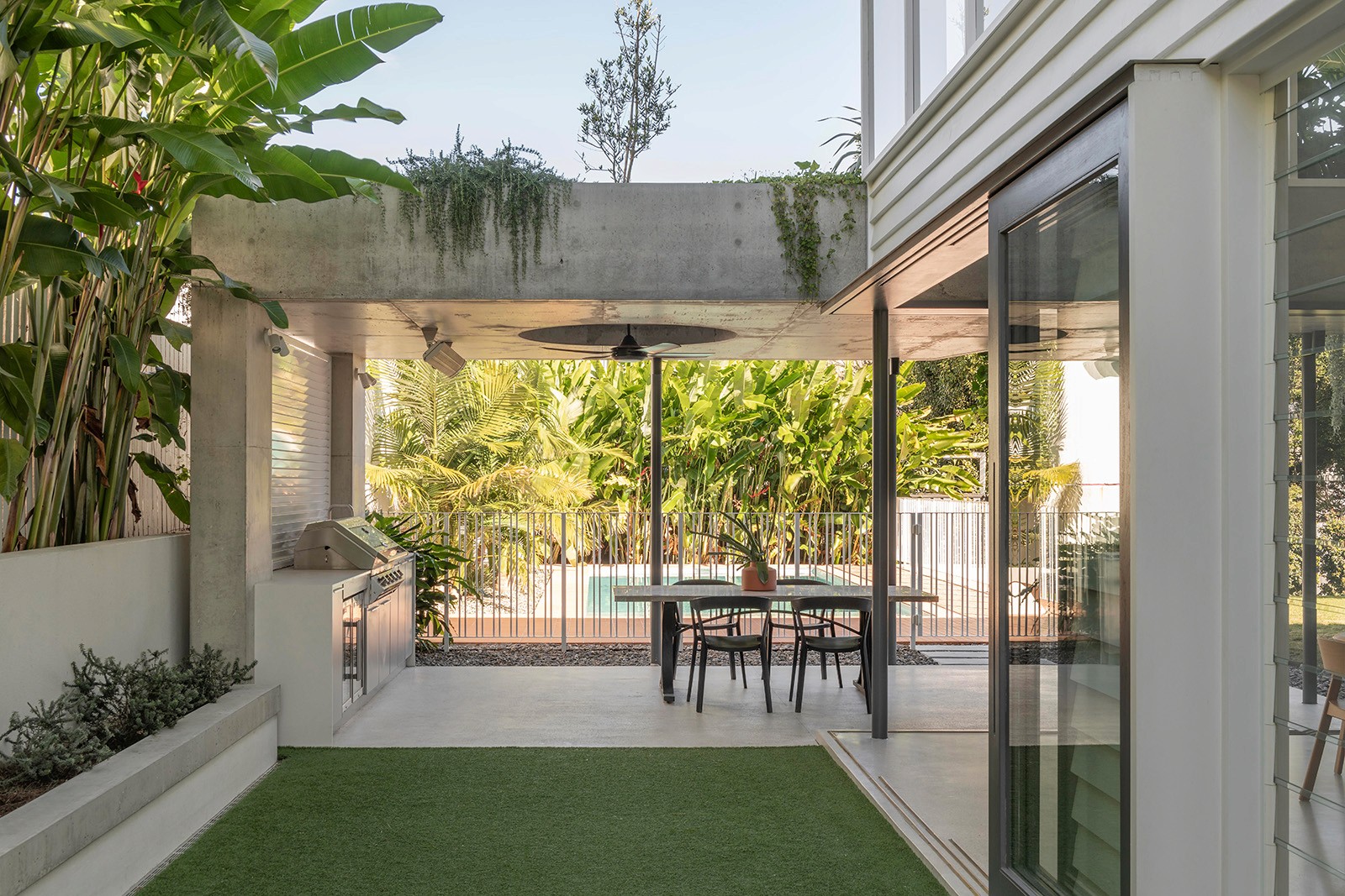 Covered outdoor dining area framed by concrete beams, lush planting, and sliding glass doors opening to the garden.