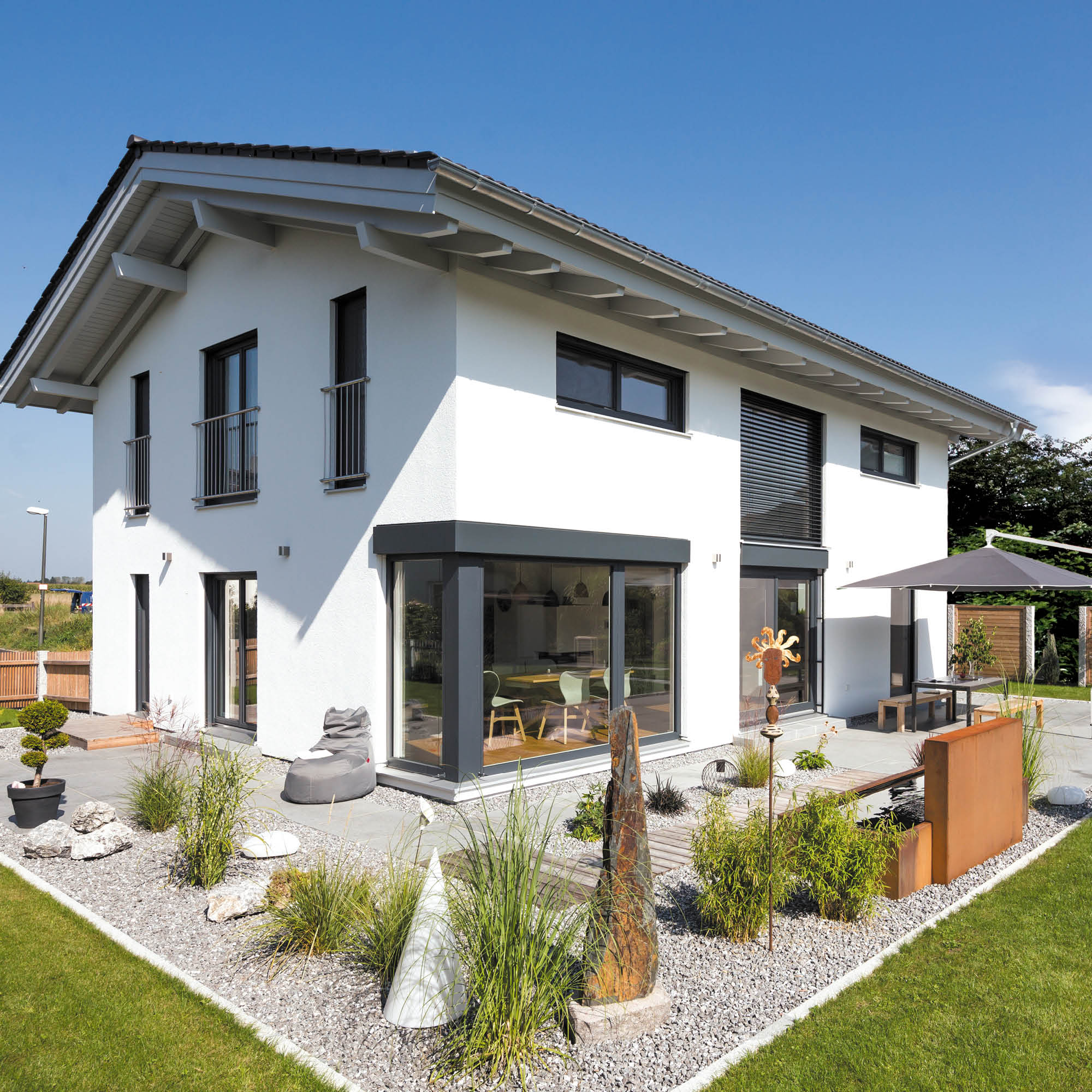 Modernes weißes Einfamilienhaus mit großem Erkerfenster, Garten mit Kieswegen, Wasserobjekt und Sitzgelegenheiten.