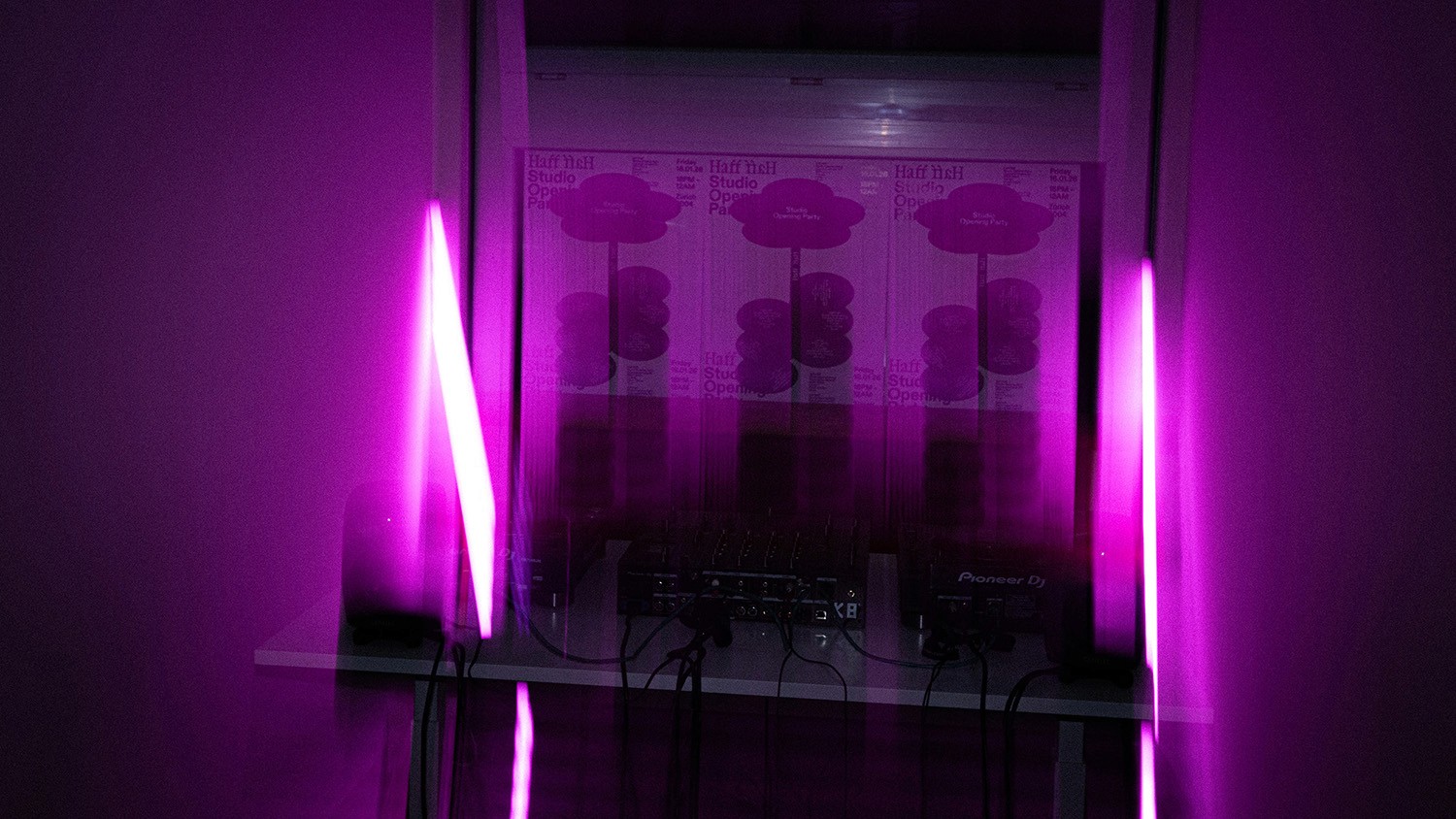 An experimental, long-exposure photograph of the DJ booth at the studio housewarming, featuring glowing pink neon light tubes and motion-blurred "Studio Opening Party" posters in the background. This artistic shot emphasizes the studio’s expertise in immersive event branding and dynamic visual storytelling.