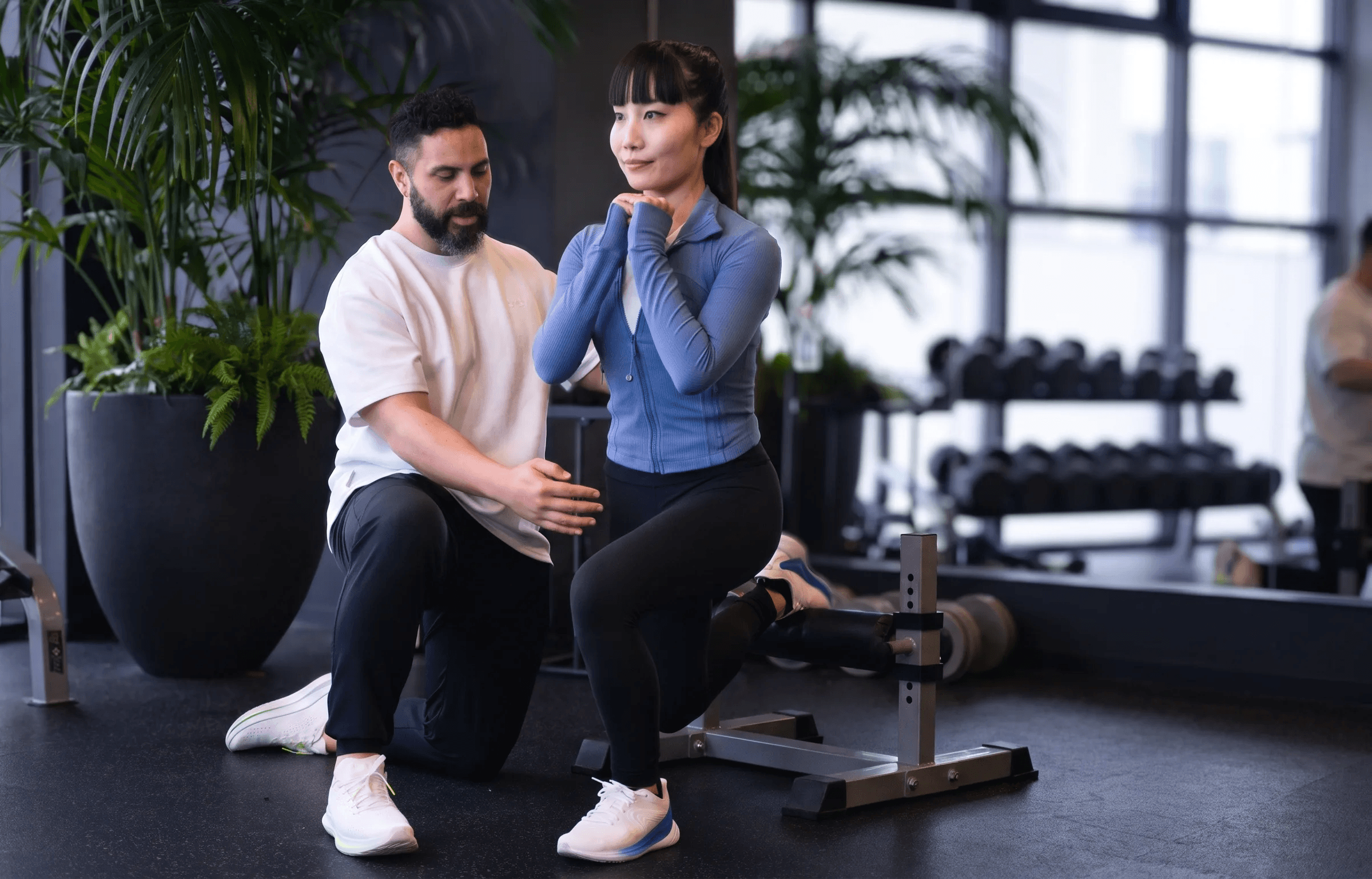A coach kneels beside a woman at the bottom of a deadlift, guiding her head and back position before she lifts