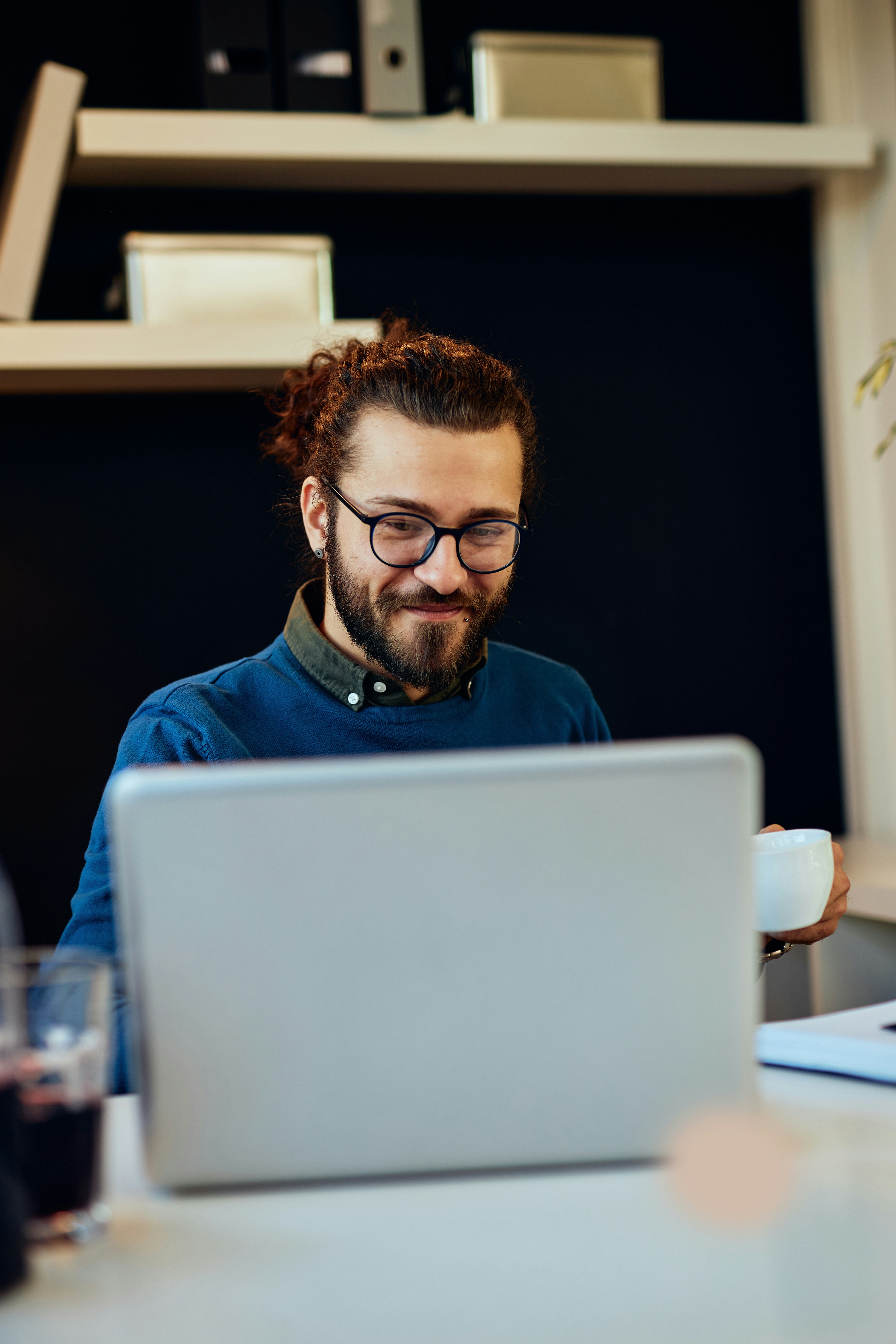 A man with glasses smiles while working on a laptop at a desk, with bookshelves in the background.