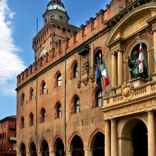 Edificio storico in mattoni con finestre ad arco, bandiere e statue sotto un cielo azzurro.