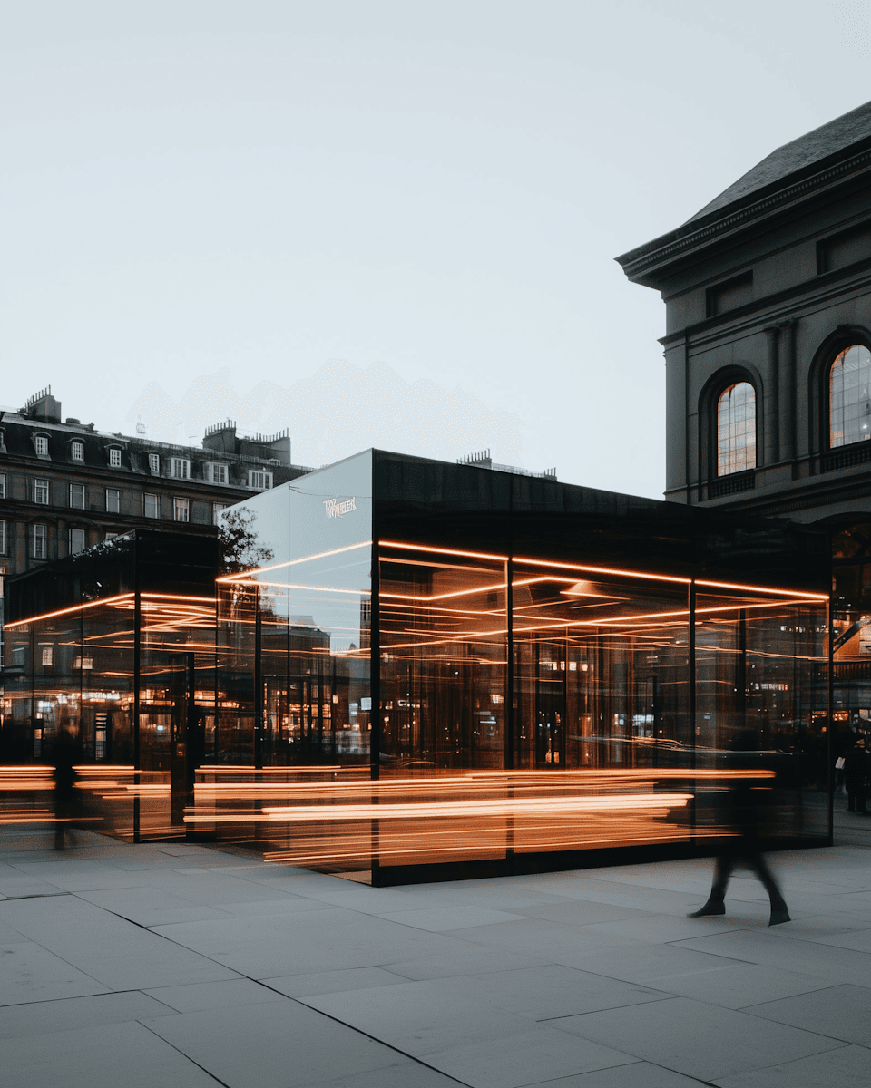 A reflective glass structure surrounded by warm lighting, set against a historic building backdrop.
