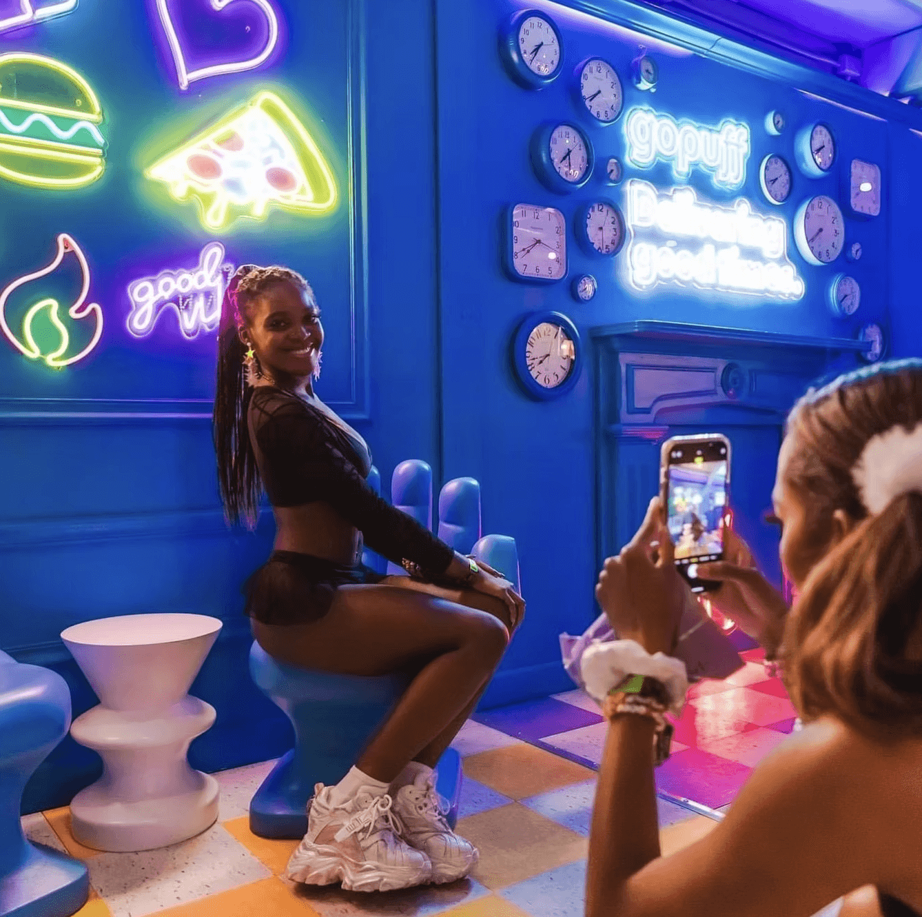 Person smiling and holding a holographic gopuff fanny pack, standing against a bright blue wall with the "gopuff / Delivering good times." neon sign and decorative clocks.