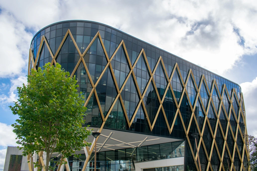 Modern multi-story building with a curved, pill-shaped footprint featuring dark reflective glass panels and a decorative gold-toned metallic lattice forming large diamond patterns across the facade. The roofline has a distinctive sawtooth edge. The recessed ground floor reveals a sheltered walkway with angular LED lighting and a visible interior with modern white furniture. A large green tree and modern street lamps stand in the foreground under a partly cloudy sky.