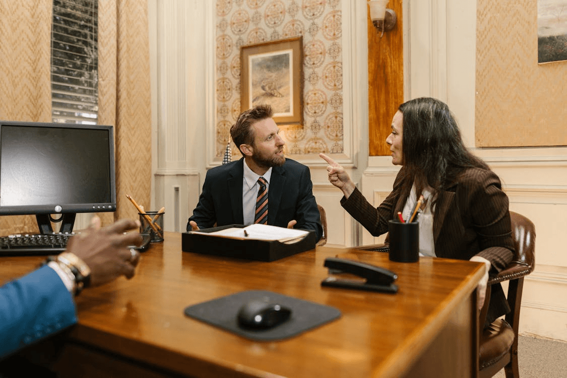A tense office meeting where two people in business attire argue across a desk, with papers and a computer monitor nearby.