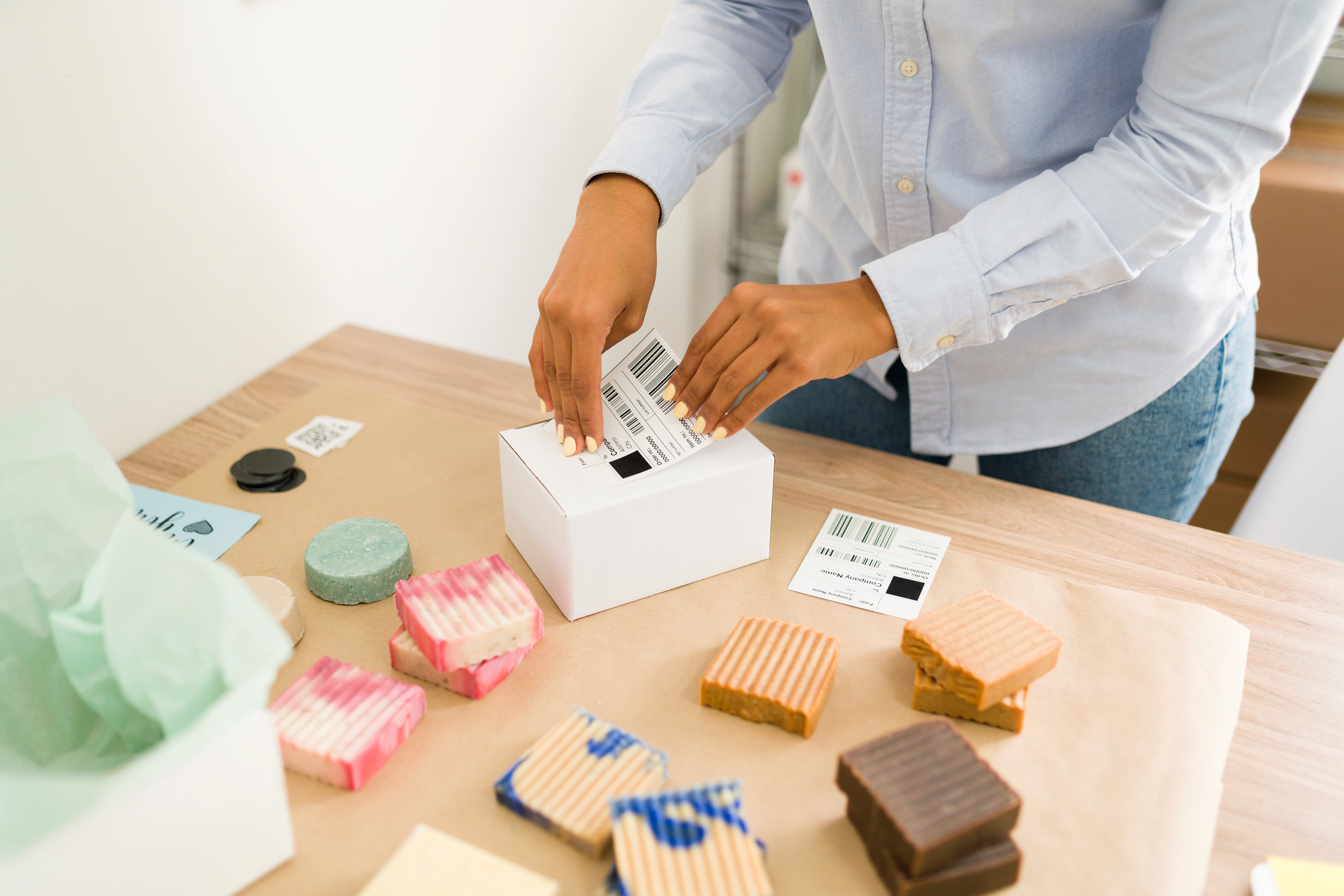 Person in a blue shirt labels a white box on a table with colorful handmade soaps. The scene conveys a sense of small business or craft packaging.