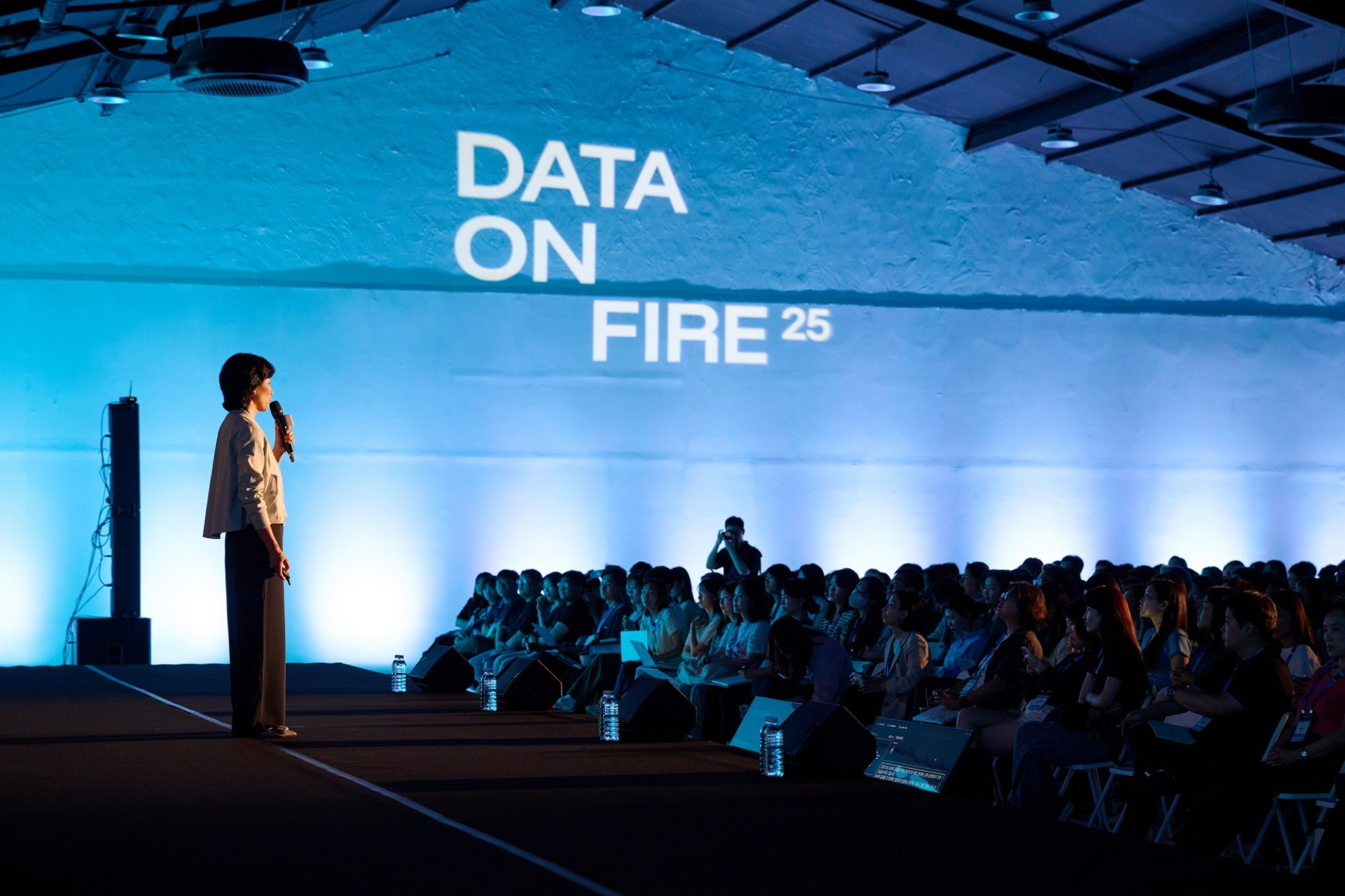 A speaker stands on stage in front of an audience, with "DATA ON FIRE" displayed on a large screen behind.