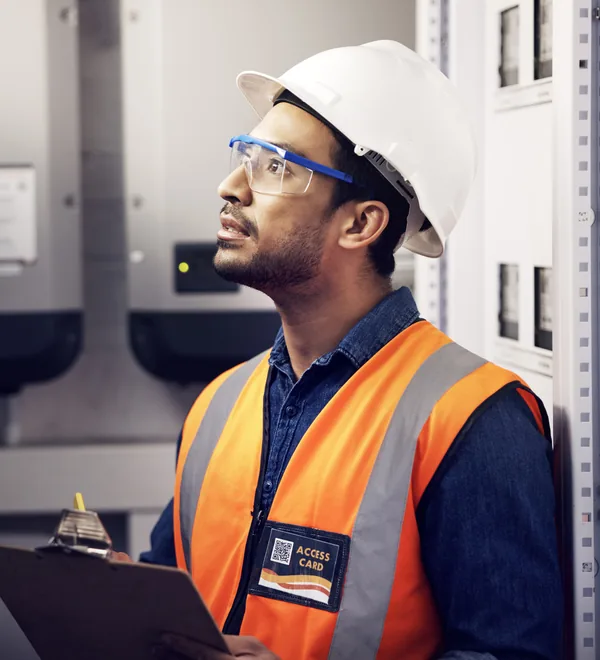 Technician with a clipboard recording readings beside electrical cabinets in a substation control room