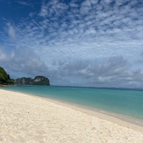 A serene beach with white sand, clear turquoise water, and a mountainous island in the distance under a partly cloudy sky.