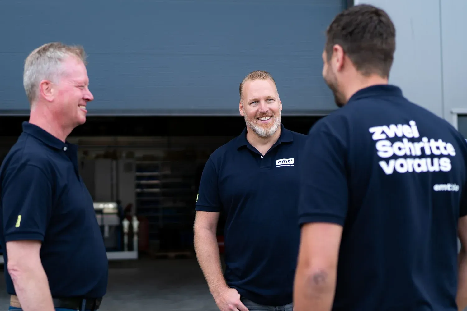 Drei Männer im Gespräch, zwei tragen Poloshirts mit "zwei Schritte voraus"-Aufdruck.