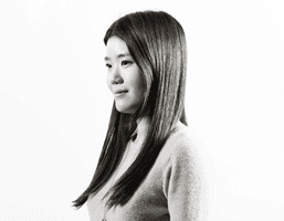 Black and white portrait of a young woman with long, straight dark hair. She is looking slightly to her left, against a plain white background. She wears a light-colored cardigan or sweater over a lighter-colored collared shirt. The image is in a portrait orientation.