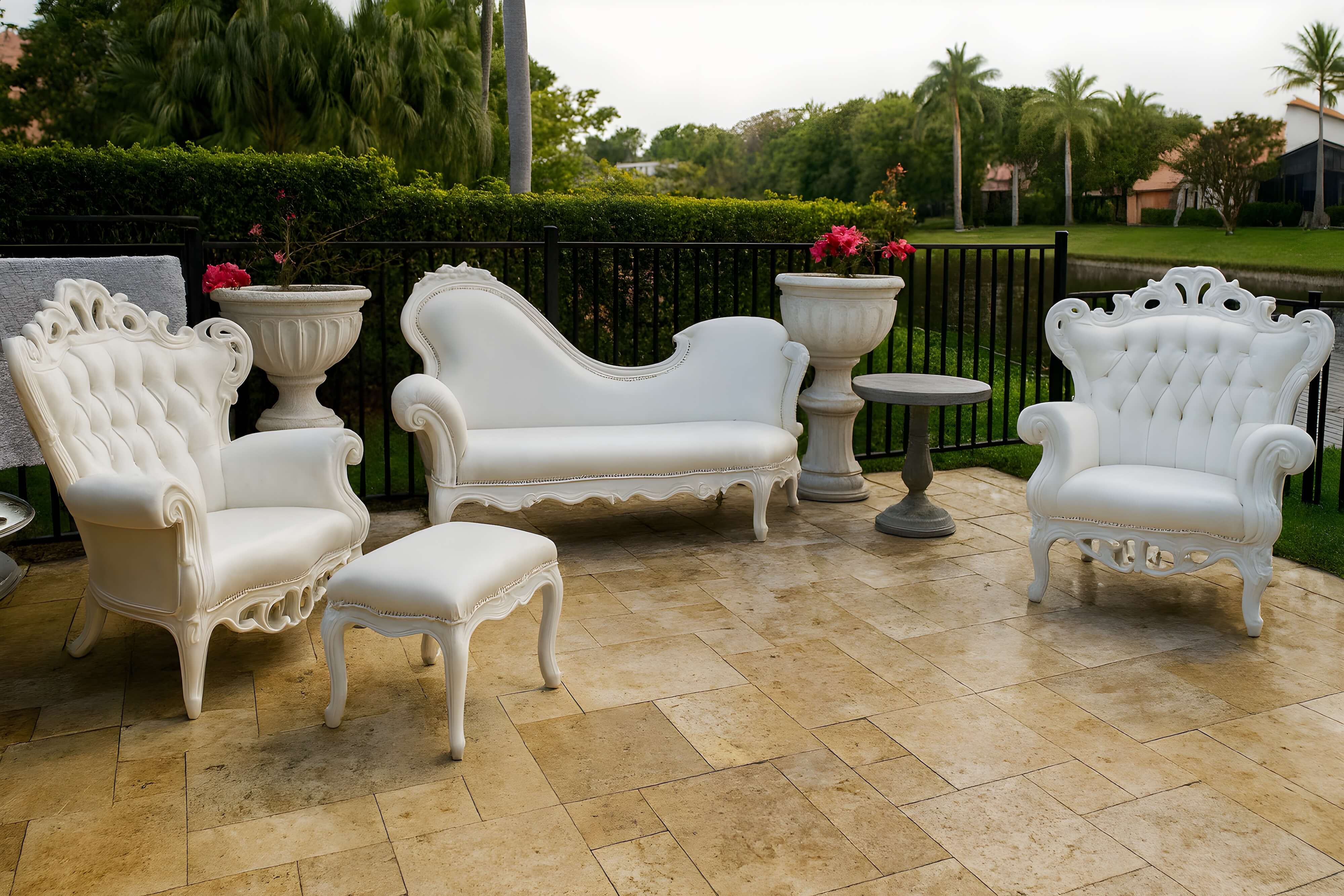 Elegant outdoor patio set featuring white tufted armchairs, a curvaceous chaise-style loveseat, matching ottoman, and stone-top side tables, set on a travertine tile terrace with lush garden and water views.