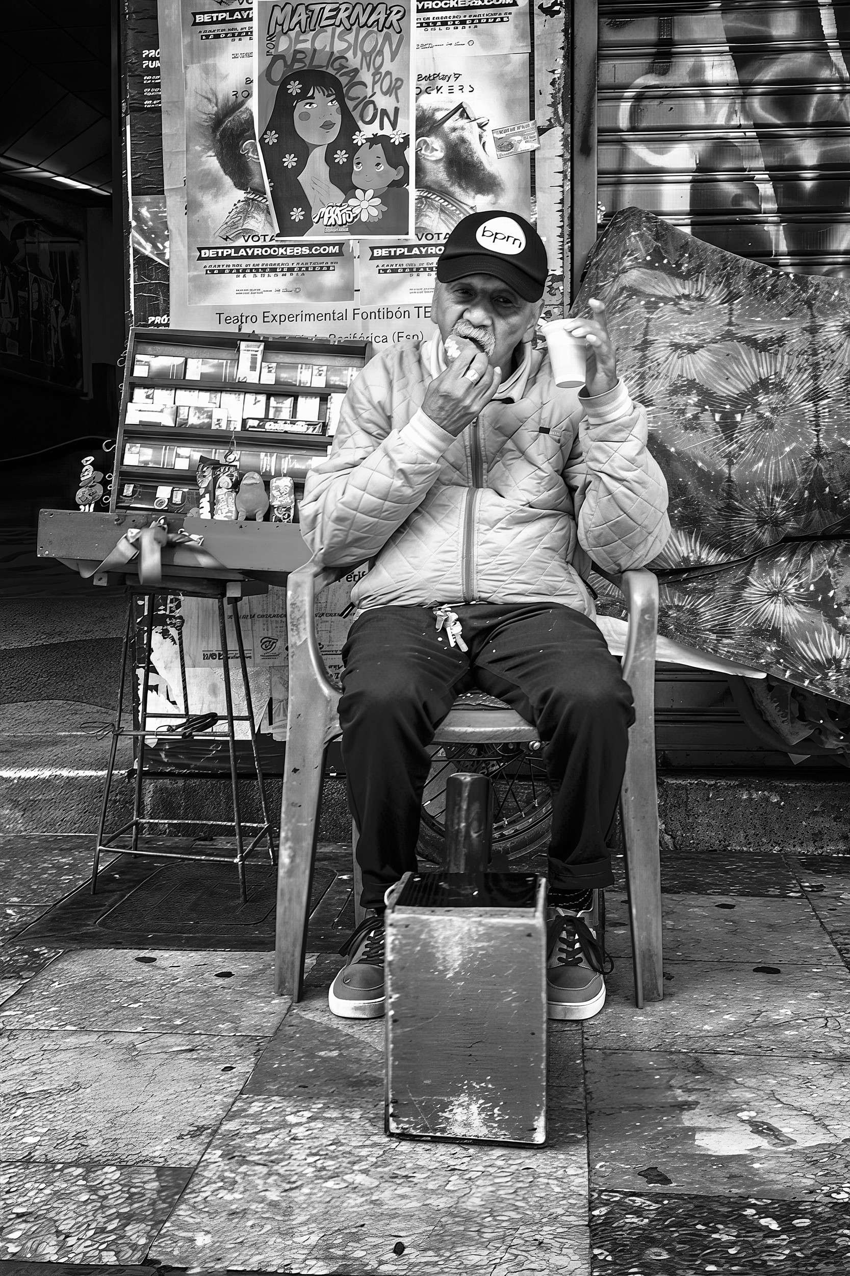 A man sits in a plastic chair on a Bogotá sidewalk, eating a snack and drinking from a paper cup, with posters and a small stall behind him.