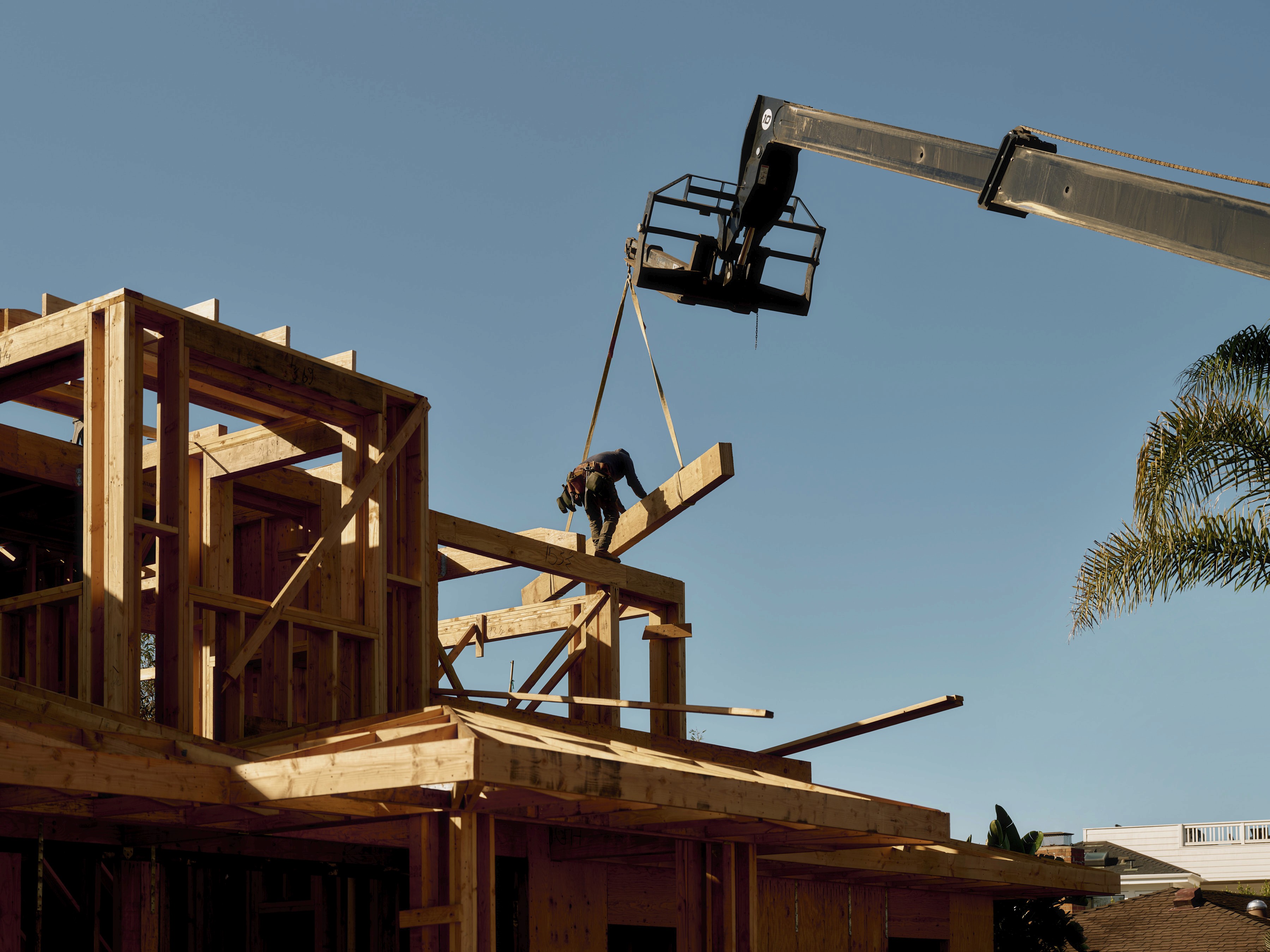 A construction worker on a wooden house frame guides a large beam being lowered by a crane against a clear blue sky.