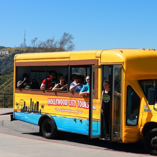 一辆黄色的好莱坞城市观光巴士，侧面开放，游客坐满座位，背景中可见好莱坞标志。