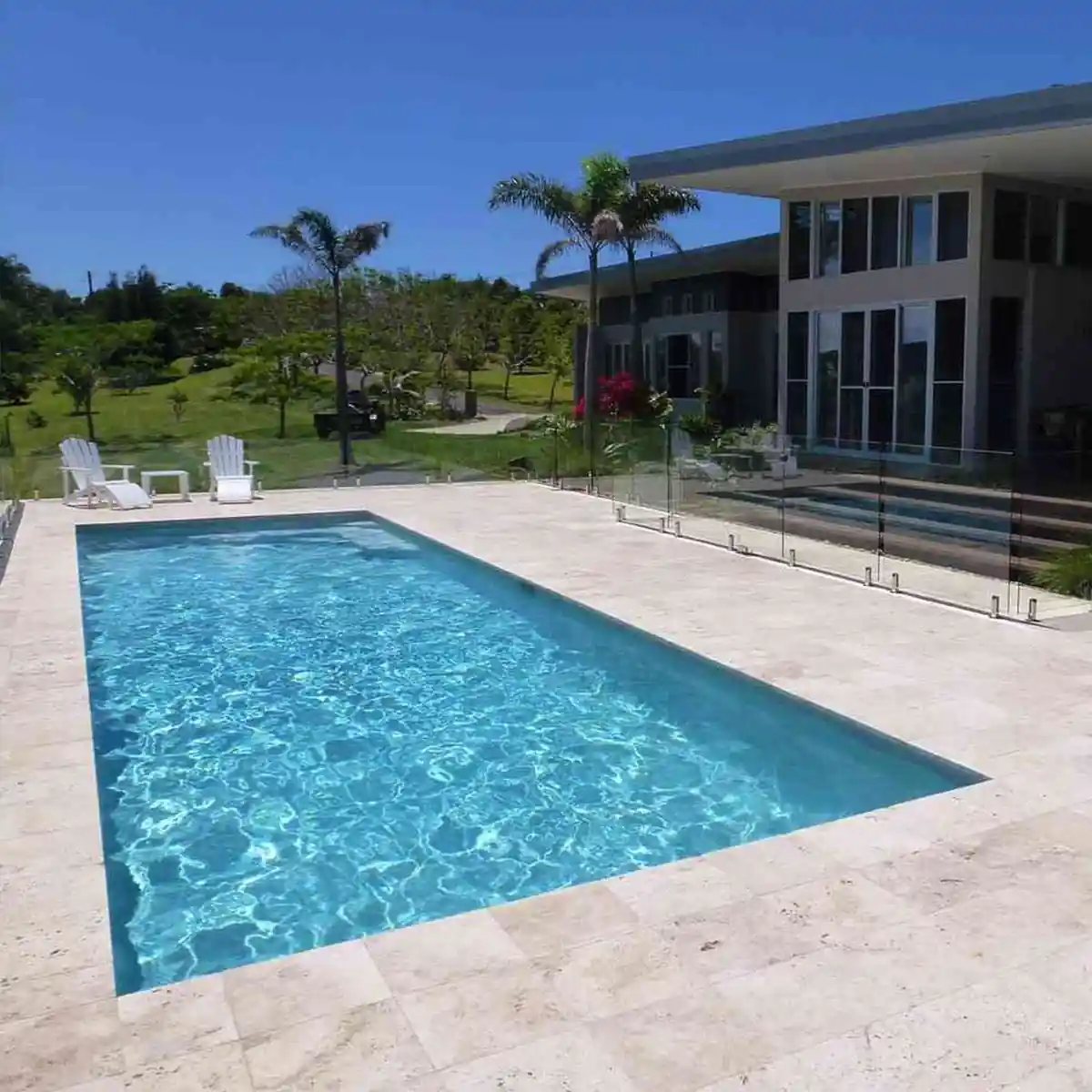 Lujosa piscina de agua turquesa con un amplio solárium perimetral de mármol travertino y cerco de vidrio de seguridad, frente a una casa moderna.