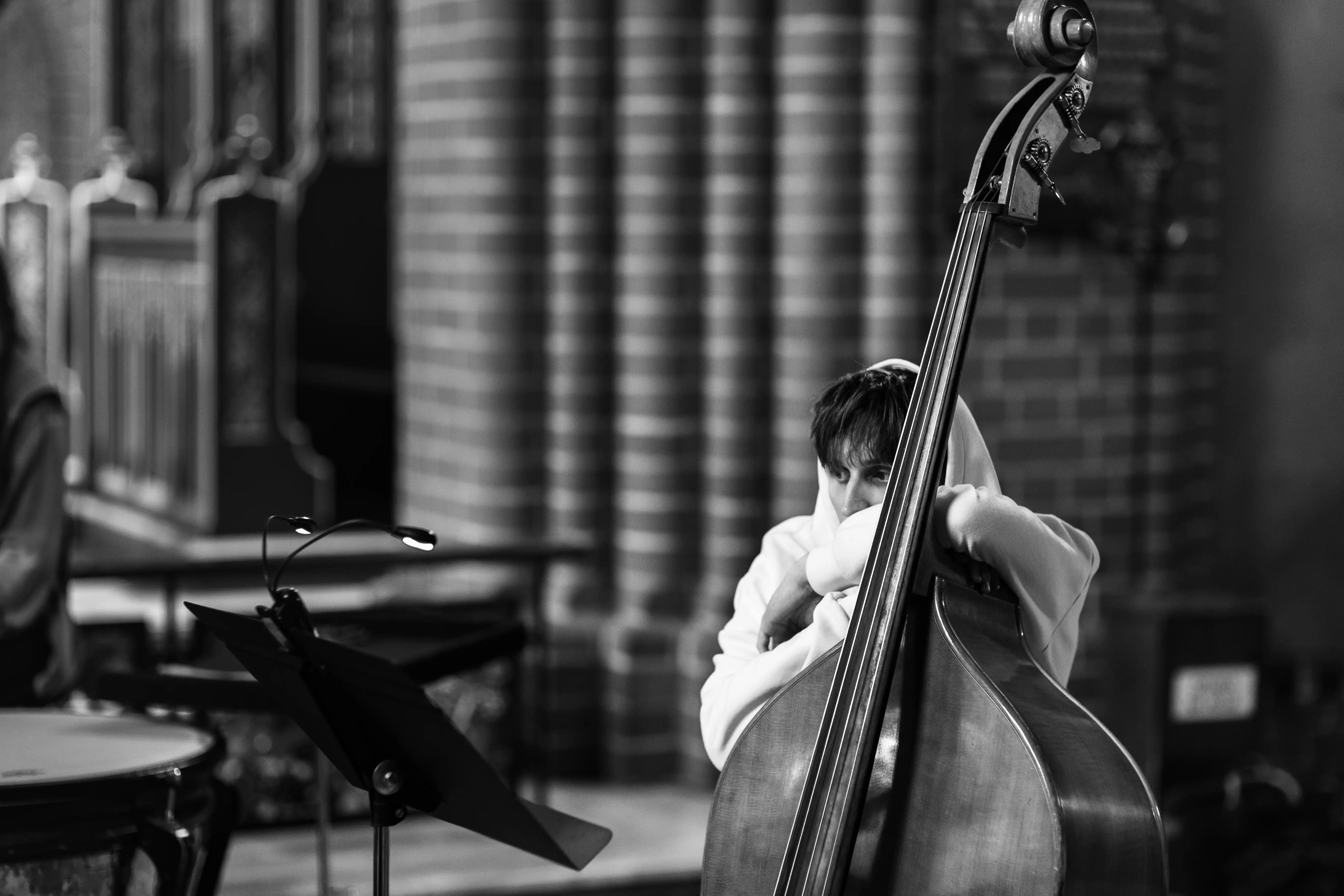 Młody muzyk grający na kontrabasie, ubrany w białą bluzę z kapturem. Ujęcie monochromatyczne przedstawia go w sali z widocznymi ceglastymi ścianami.