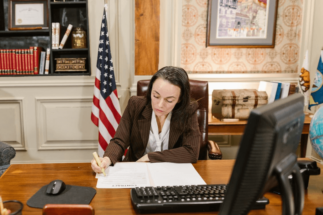 A woman in a suit reviewing and signing a document at an office desk, with an American flag behind her.
