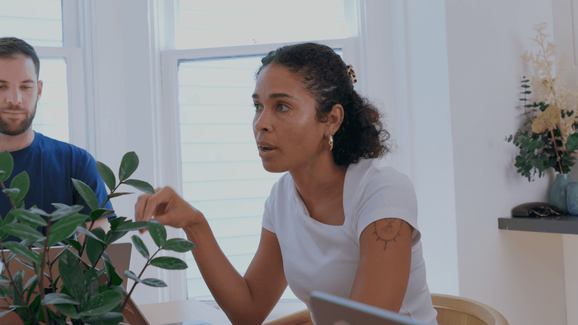 A person with short black hair and glasses smiles softly, wearing a black shirt against a dark background with a soft light illuminating one side, creating a warm and inviting atmosphere.