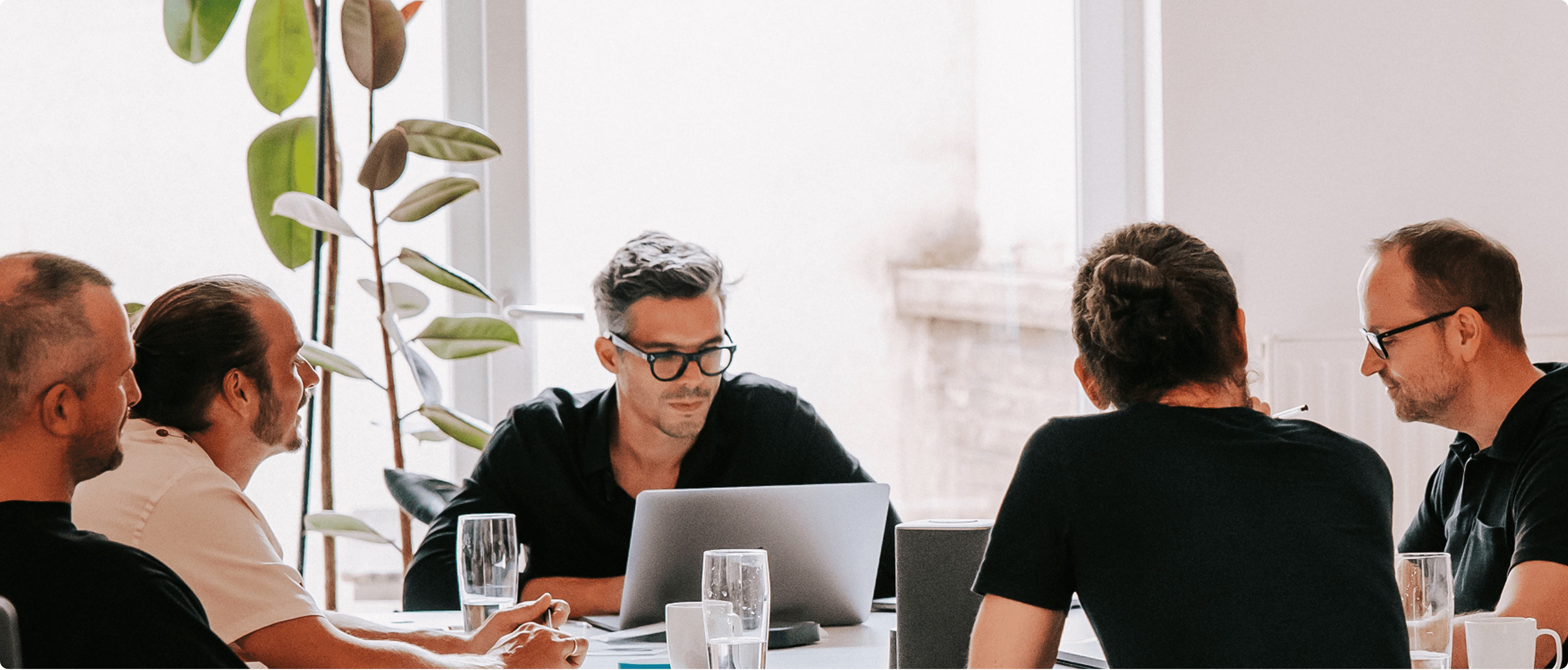 Team members gathered around a table in a bright office, collaborating during a workshop with laptops.