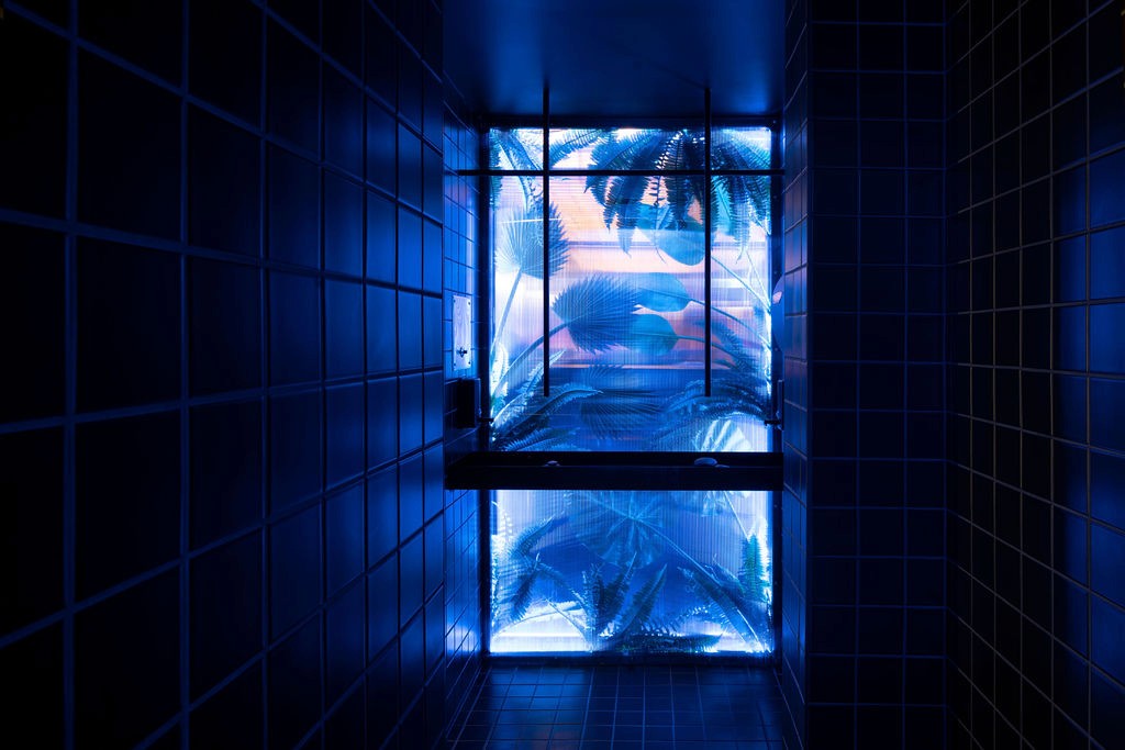 Futuristic washroom design at Minéral, featuring glowing blue light, reflective surfaces, and lush greenery framed by a geometric grid of tiles.