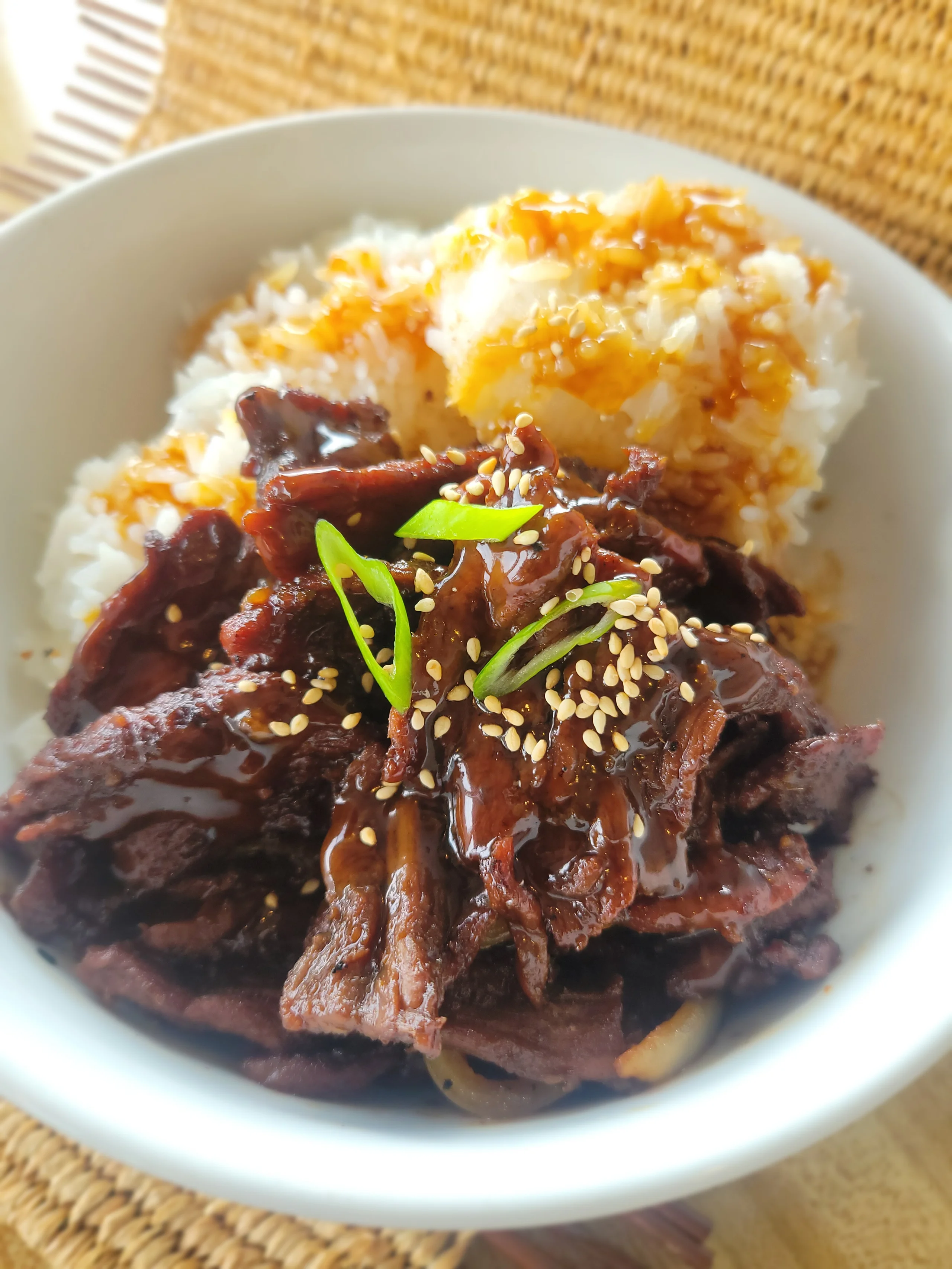 A white bowl filled with tender, glazed slices of teriyaki beef topped with sesame seeds and green onions, served over three scoops of white rice.