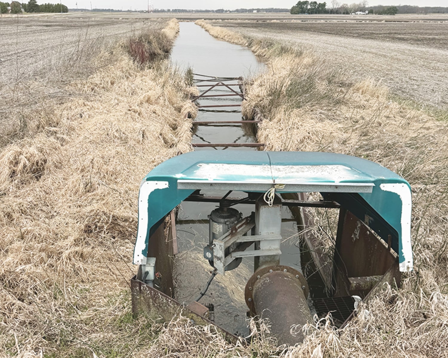 Water pump structure with turquoise cover, set beside a drainage canal lined with dry grass.