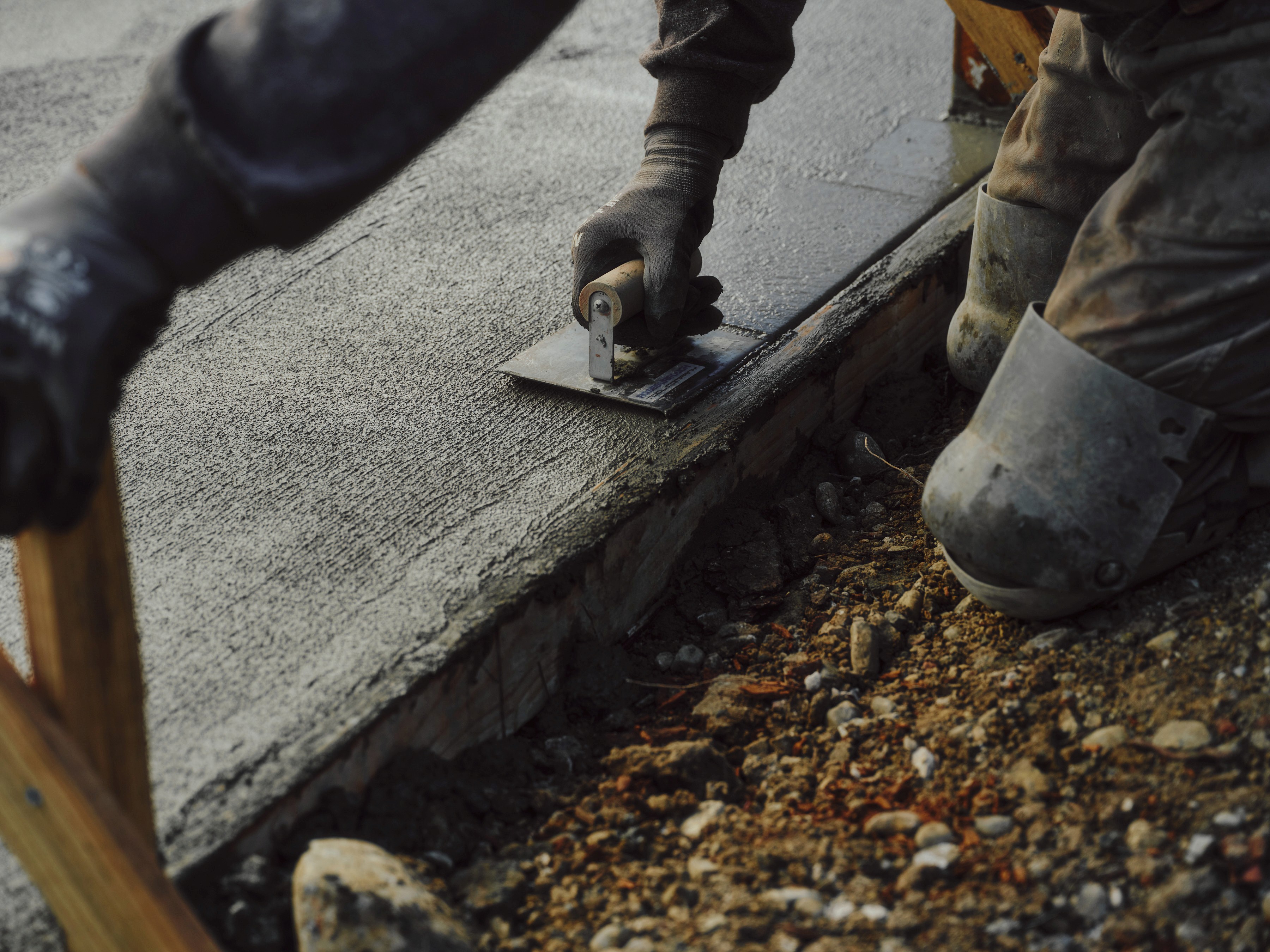 Close-up of a mason finishing concrete with a metal trowel along a wooden form, wearing protective work gloves and heavy-duty knee pads