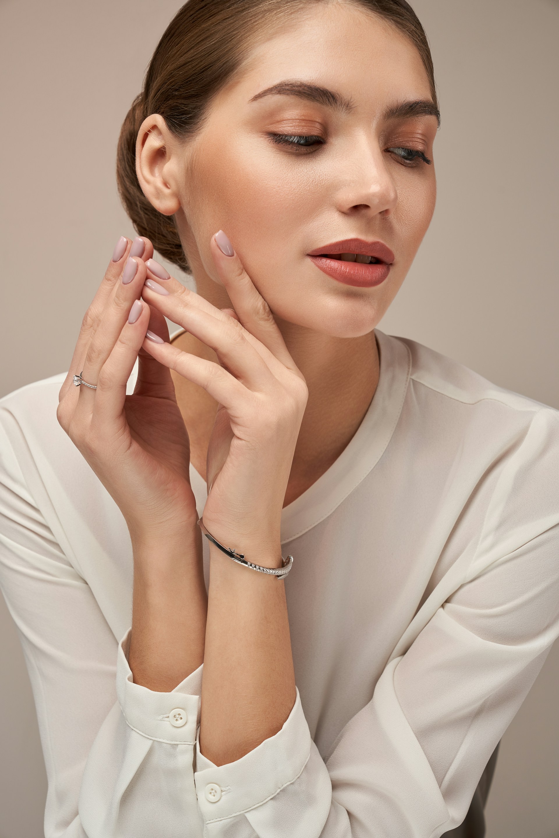 A woman with her hair pulled back in a sleek bun, wearing a white blouse and delicate silver jewelry, with her hands raised near her face.