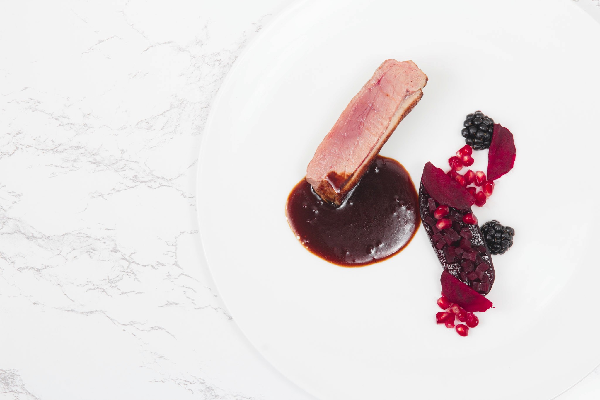 Duck breast served with a pool of blackberry sauce, glazed beetroot, blackberries, and pomegranate on a white plate