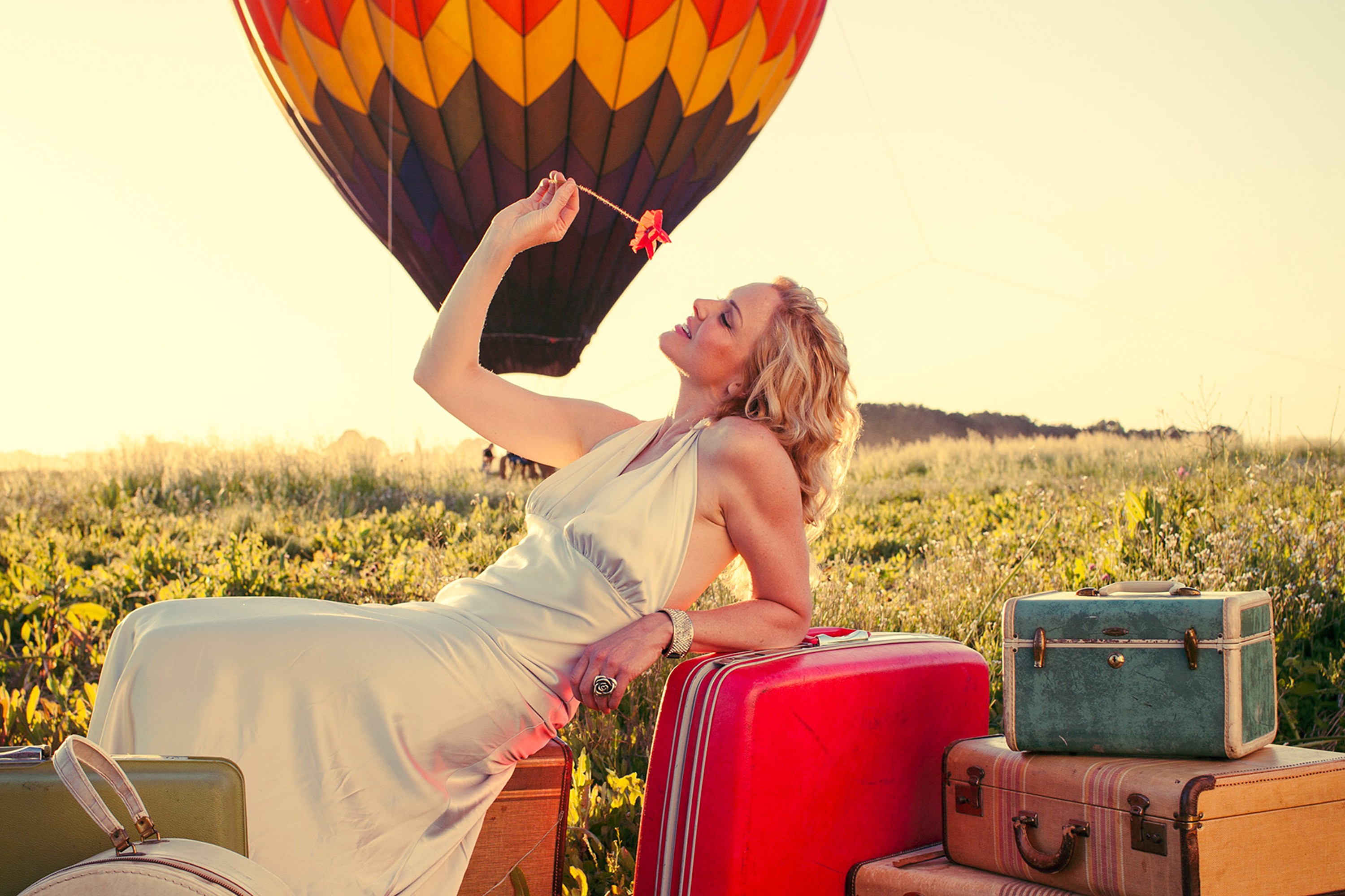 A 30s white woman laying on a pile of suitcases in middle of a field while there is a hot air balloon in the background.