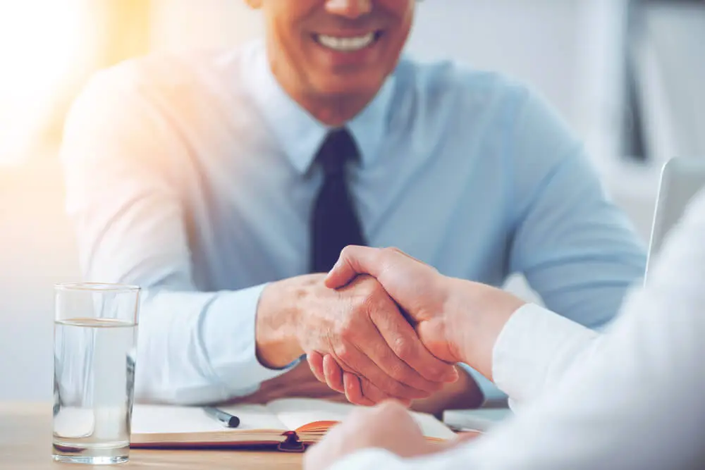 Two people shaking hands during a business meeting, with one smiling man in a shirt and tie visible