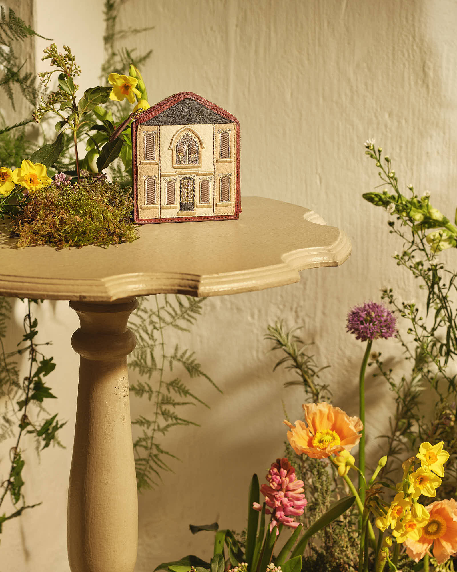 Elegant small house model with intricate façade on table surrounded by vibrant flowers.
