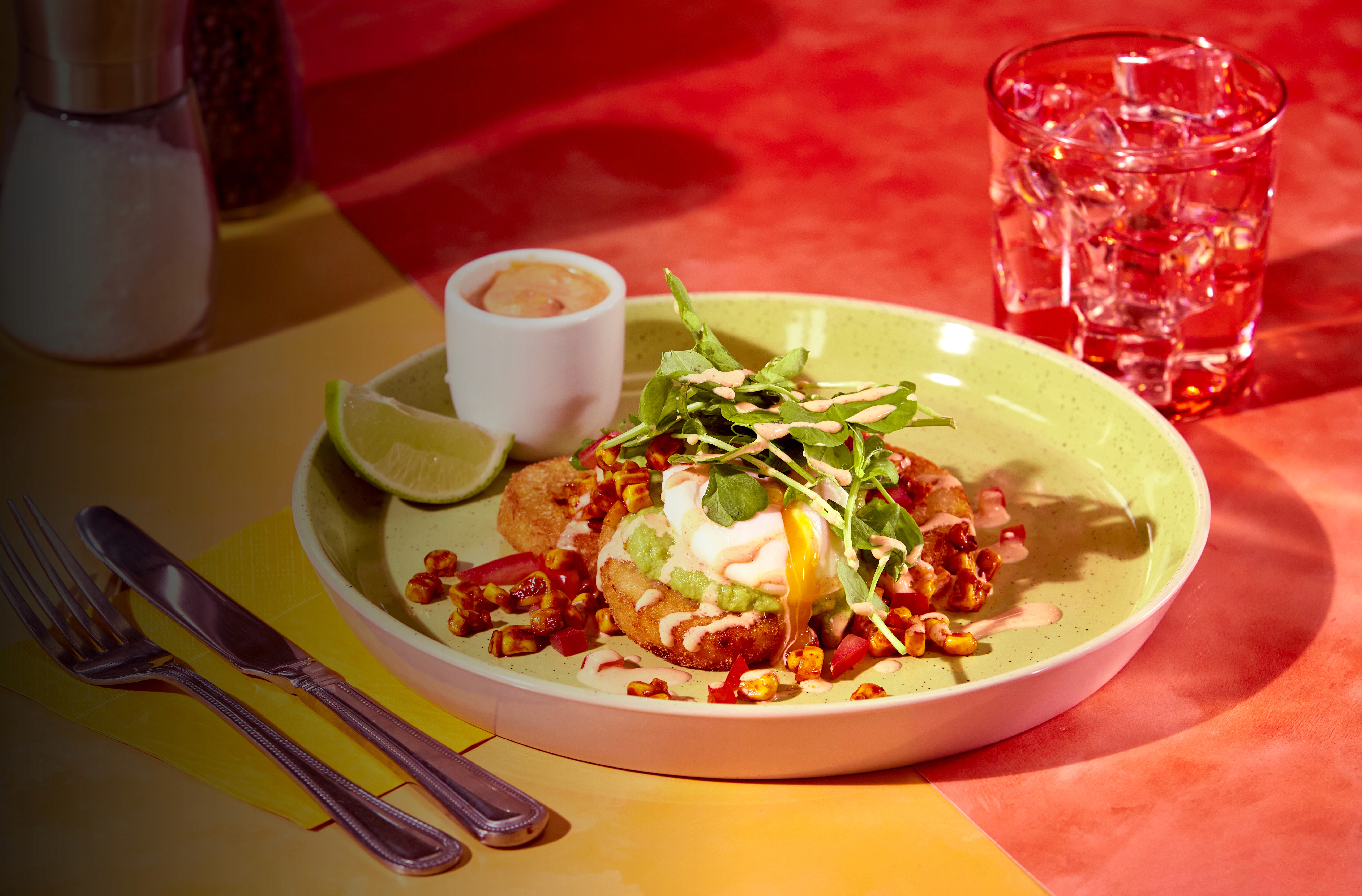 A colourful plated dish featuring a Strong Roots cauliflower hash brown, topped with a poached egg, avocado, microgreens, and creamy sauce, garnished with roasted corn and diced red peppers. Served with a lime wedge and a small cup of sauce on the side, alongside a glass of water, cutlery, and salt and pepper shakers on a vibrant red and yellow table