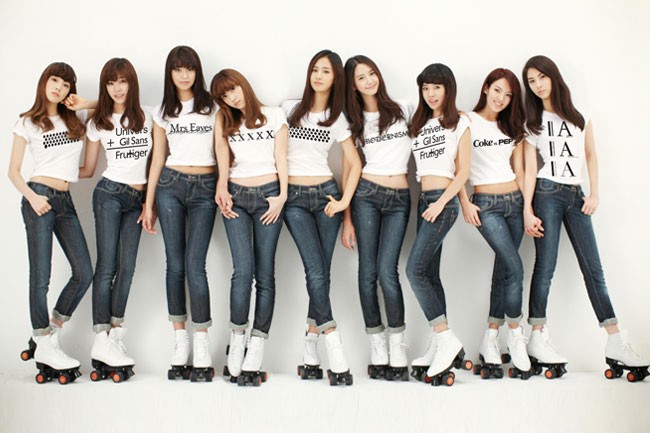 Ten young women stand in a row against a white wall, wearing white t-shirts with black text and blue jeans. Each shirt displays a unique text phrase: “Universe + Gil Gans Frutger,” “Mrs.Eaves,” “Mrs.Eaves,” “Mrs.Eaves,” “Mrs.Eaves,” “Mrs.Eaves,” “Mrs.Eaves,” “Mrs.Eaves,” and “Mrs.Eaves.” The women are wearing white sneakers and roller skates.